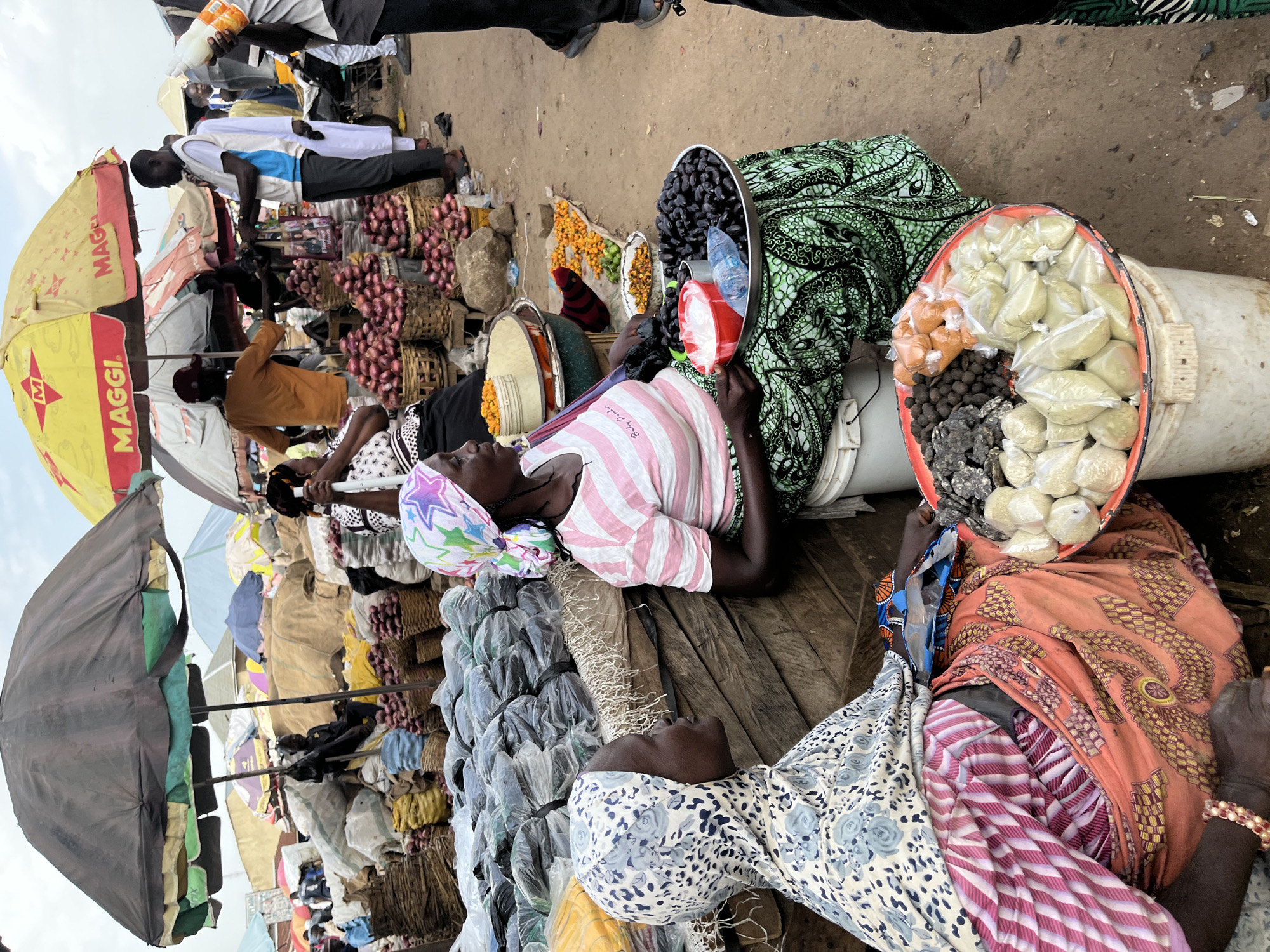 In Rafin Par, Nigeria, a bustling outdoor market scene unfolds under a cloudy sky. Two women, dressed in vibrant headscarves and patterned clothing, sit amidst their wares. The woman on the left wears a blue and white floral headscarf and a pink and white striped shirt, her left hand resting on her lap adorned with a beaded bracelet. The woman on the right, her headscarf a colorful mix of pink, purple, and green stars, is also in a pink and white striped shirt.
Their display features an array of goods on a wooden surface. To the left, a woman is partially visible behind sacks of produce. In the center, a large metal tray holds dark, round fruits, possibly dates or olives, alongside two red cups and a plastic bottle of water. A second, smaller red tray sits in front of the seated women, piled with various items. These include dark, lumpy spices or dried fruits, small orange powder-filled packets, and numerous small plastic bags filled with a light green powder or dried herbs.
The market is filled with other vendors and customers, many under colorful umbrellas, including a prominent yellow Maggi-branded umbrella. Baskets overflowing with red onions are visible in the background, as are large sacks of produce and more packaged goods. The ground is dusty earth, characteristic of a market in this region. The overall impression is one of lively commerce and community in a Nigerian marketplace.