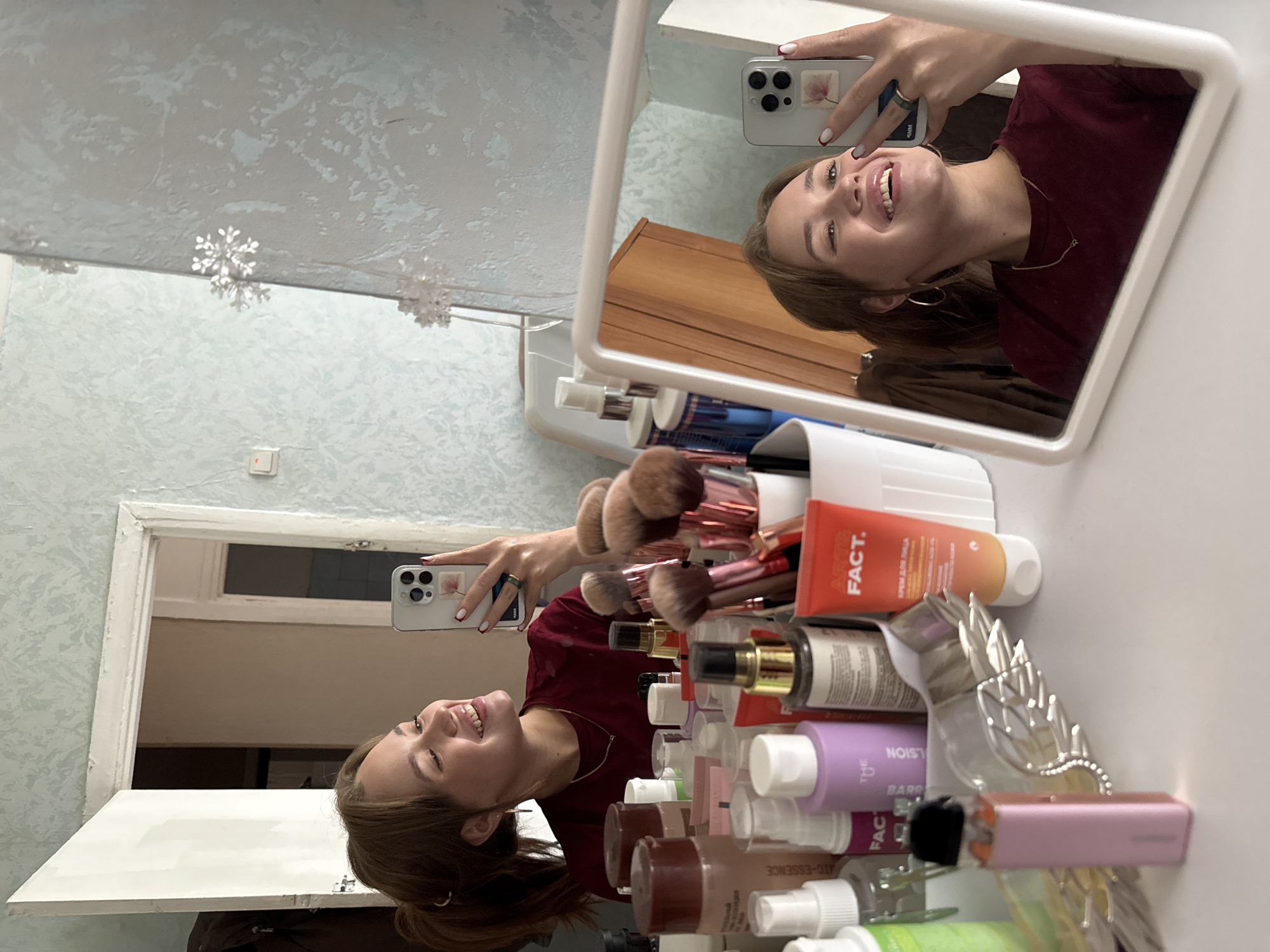 The image shows a young woman taking a selfie in a mirror. She is wearing a dark red t-shirt and large hoop earrings, with her hair down. Her face is illuminated, and she appears to be smiling or laughing. In her right hand, she holds a smartphone, which is capturing the reflection. The background of the mirror's reflection shows a cluttered vanity with various cosmetic items, including makeup brushes, tubes, and bottles. One tube is orange with the brand "FACT." visible. In the foreground, the woman's hand is visible holding the phone, with a ring on her left ring finger and manicured nails.
The scene is indoors, likely in a bathroom or dressing area, given the presence of makeup and the mirror. The lighting suggests it is daytime. The overall impression is one of casualness and self-expression. The wall texture in the reflection appears to be a light blue patterned wallpaper. Snowflake decorations are visible on the wall and the ceiling. The woman is looking directly at the camera in the reflection, with a playful expression.
