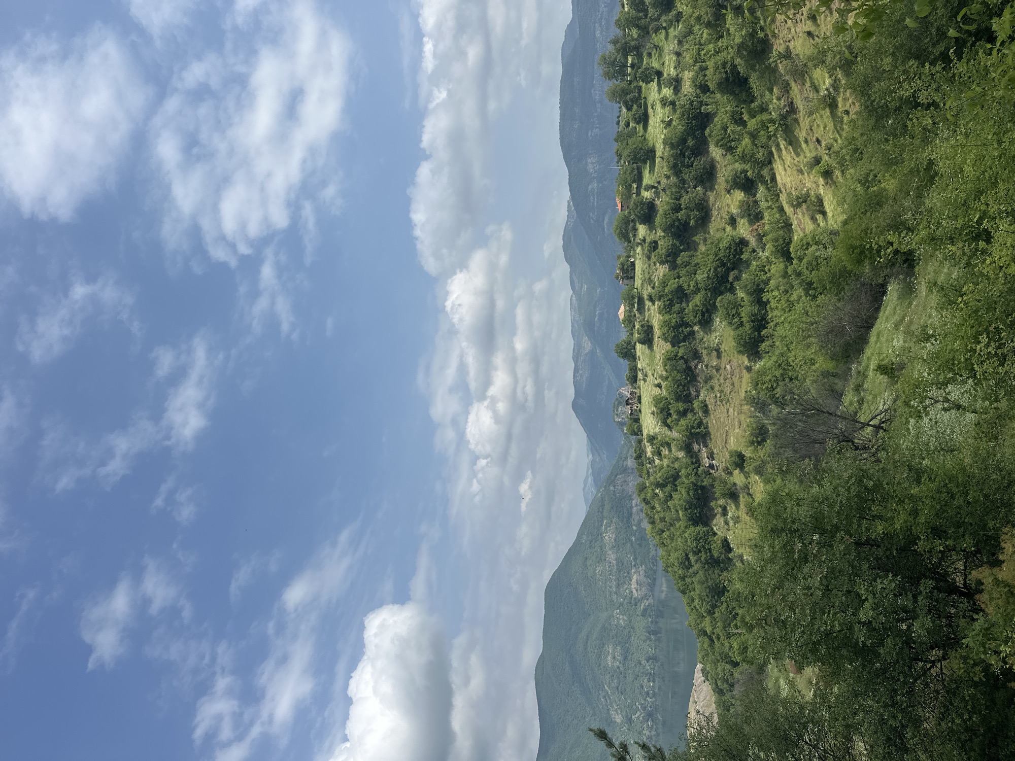 A wide landscape view depicts a serene mountainous region under a bright, partly cloudy sky. The upper portion of the scene features a clear blue sky dotted with scattered white cumulus clouds, varying from wispy to fluffy in appearance.

Below the sky, rolling green hills and mountains dominate the landscape. In the mid-ground, a large hillside is covered with dense, vibrant green vegetation, including numerous trees and patches of sunlit grass. Several small structures with reddish-orange roofs are visible on this hillside, suggesting scattered dwellings. Further into the distance, successive layers of darker, more rugged mountains stretch across the horizon, partially obscured by a light atmospheric haze. A body of water, possibly a lake or river, can be seen nestled within the valley, reflecting the sky and distant terrain.

The foreground, particularly towards the right, is filled with dark green foliage from trees and bushes, indicating the photograph was taken from an elevated vantage point overlooking a valley or gorge. The overall impression is one of natural beauty and tranquility, typical of a rural, mountainous environment. The lighting suggests it is daytime, likely late morning or early afternoon, with pleasant weather conditions. The setting is Star Chitak, Bulgaria. No people or discernible activities are present, and no text is visible in the image.