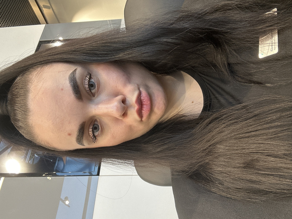 A female individual is depicted in a chest-up portrait, seated in an indoor, modern environment. She has long, dark brown hair styled in a high ponytail, with loose strands cascading over her shoulders. Her eyes are light blue, and her eyebrows are dark and well-defined. A small red blemish is present on her forehead. She is looking directly at the camera with her lips intentionally pursed. She is wearing a dark, possibly black, short-sleeved, crew-neck top. A white rectangular name tag is affixed to her left side, bearing Cyrillic text that identifies her as "София Дро..." (Sofia Dro...) and her role as "Администратор" (Administrator). The background shows the dark backrest of a chair, a light-colored wall or partition to the viewer's right, and a dark, reflective panel to the viewer's left, above which a white grid ceiling structure is visible. The scene is located in Saint Petersburg, Russia, within an administrative or commercial establishment.