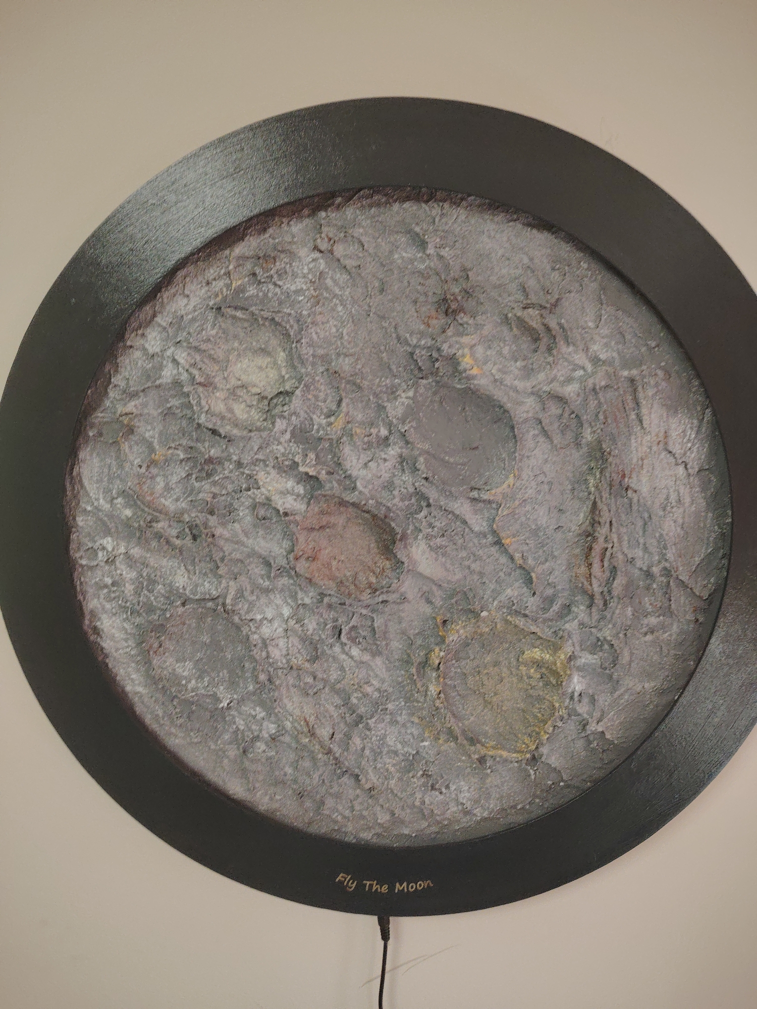 An eye-level shot captures a circular, textured artwork resembling the moon, mounted on a plain, light-colored wall, likely indoors in Huntingdonshire, United Kingdom. The artwork features a thick, dark grey or black circular frame enclosing a highly detailed, relief-like surface. This interior surface is heavily textured, simulating a lunar landscape with visible craters, ridges, and valleys.
The color palette of the moon-like surface consists predominantly of varying shades of grey, from dark charcoal to light ash, with subtle hints of reddish-brown, pale green, and dull yellow tones interspersed within the textures. These color variations contribute to the realistic, rocky appearance of the celestial body. The rough, uneven texture gives a strong sense of depth and three-dimensionality.
At the bottom center of the dark frame, the words "Fly The Moon" are inscribed in a light, possibly gold, font. A thin black wire extends downwards from the bottom edge of the frame, suggesting the artwork may be illuminated or require a power source. The surrounding wall is a uniform, light beige or off-white, providing a neutral backdrop that accentuates the circular art piece. The composition is static, focusing entirely on the artwork itself.