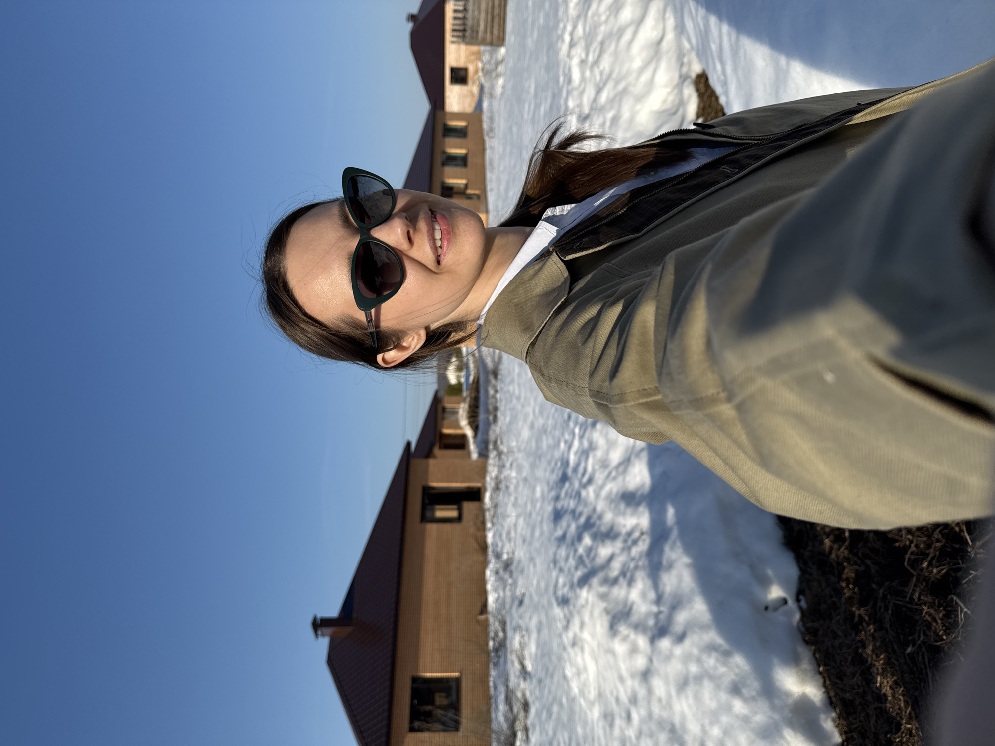 A female individual is depicted in a selfie, positioned outdoors in what appears to be a developing residential area in Chaikovsky, Russia. She has dark hair, wears dark green cat-eye sunglasses, an olive green jacket, and a light-colored collared shirt. Her head is slightly tilted, and she is smiling. The background features several single-story houses with light brown brick exteriors and dark reddish-brown gabled roofs, some appearing to be under construction or recently built. The ground is partially covered with patches of melting snow, revealing dark earth and sparse, dry vegetation. The sky above is clear and blue, indicating a bright, sunny day. No specific street or landmark is discernible.