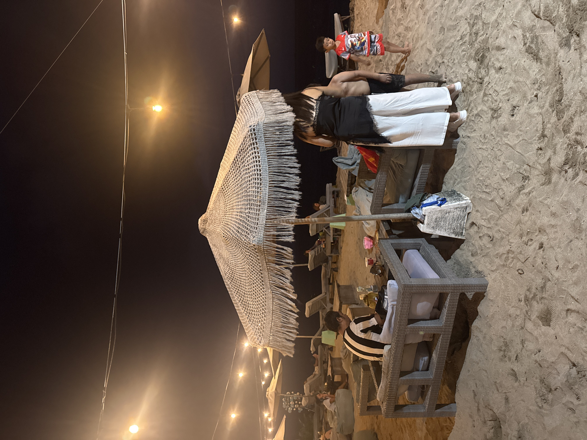 The image depicts an outdoor scene at night, likely on a beach in Denpasar, Indonesia, given the presence of sand and beach umbrellas. Several people are present, including one wearing a black and white striped shirt sitting in a gray wicker chair and another person wearing a black top and white pants. A child in a red shirt is also present. The scene is illuminated by string lights and artificial lighting, casting shadows on the sand. There are multiple beach umbrellas, some with fringed edges, and beach lounge chairs scattered around. Various objects such as drinks, bottles and towels can be seen on and around the tables and chairs. The environment is primarily sandy with a dark night sky in the background.