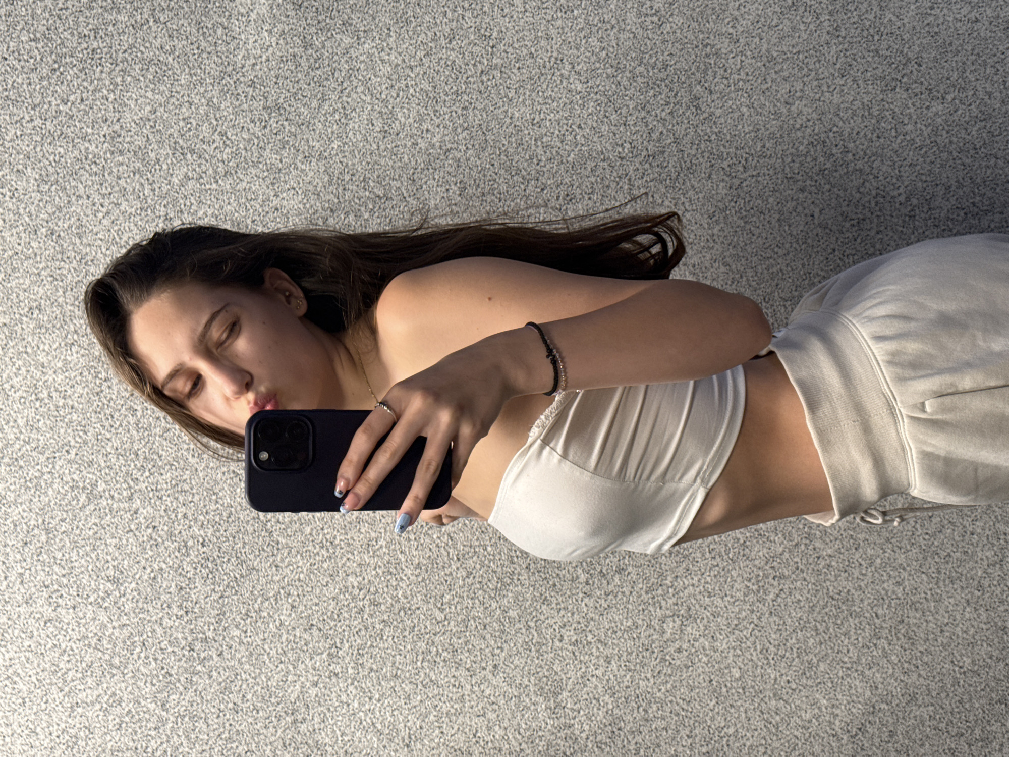 A young woman with long brown hair is taking a mirror selfie against a textured gray wall. She is wearing a white strapless top and light-colored sweatpants. Her left arm is raised, holding a dark-colored smartphone, and her lips are pursed as if blowing a kiss. She is wearing a ring on her right hand and a bracelet on her left wrist. The background appears to be an indoor setting with a speckled gray wall. There is no indication of the city of Sofia, Bulgaria.
