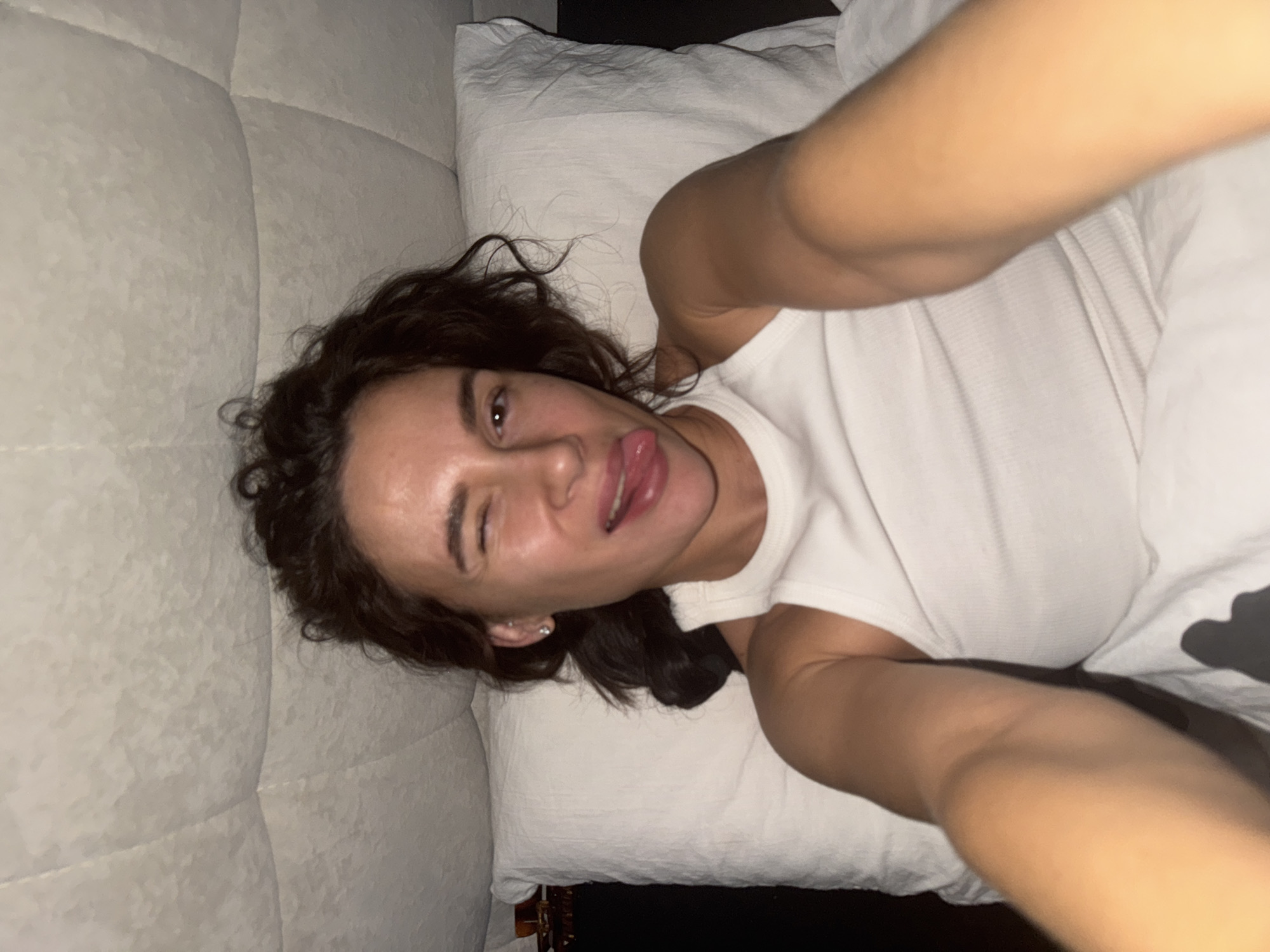 A person with brown hair and a white tank top is lying on a bed with a white pillow and headboard. The person is sticking their tongue out and winking at the camera. The location is unknown and cannot be determined from the image.