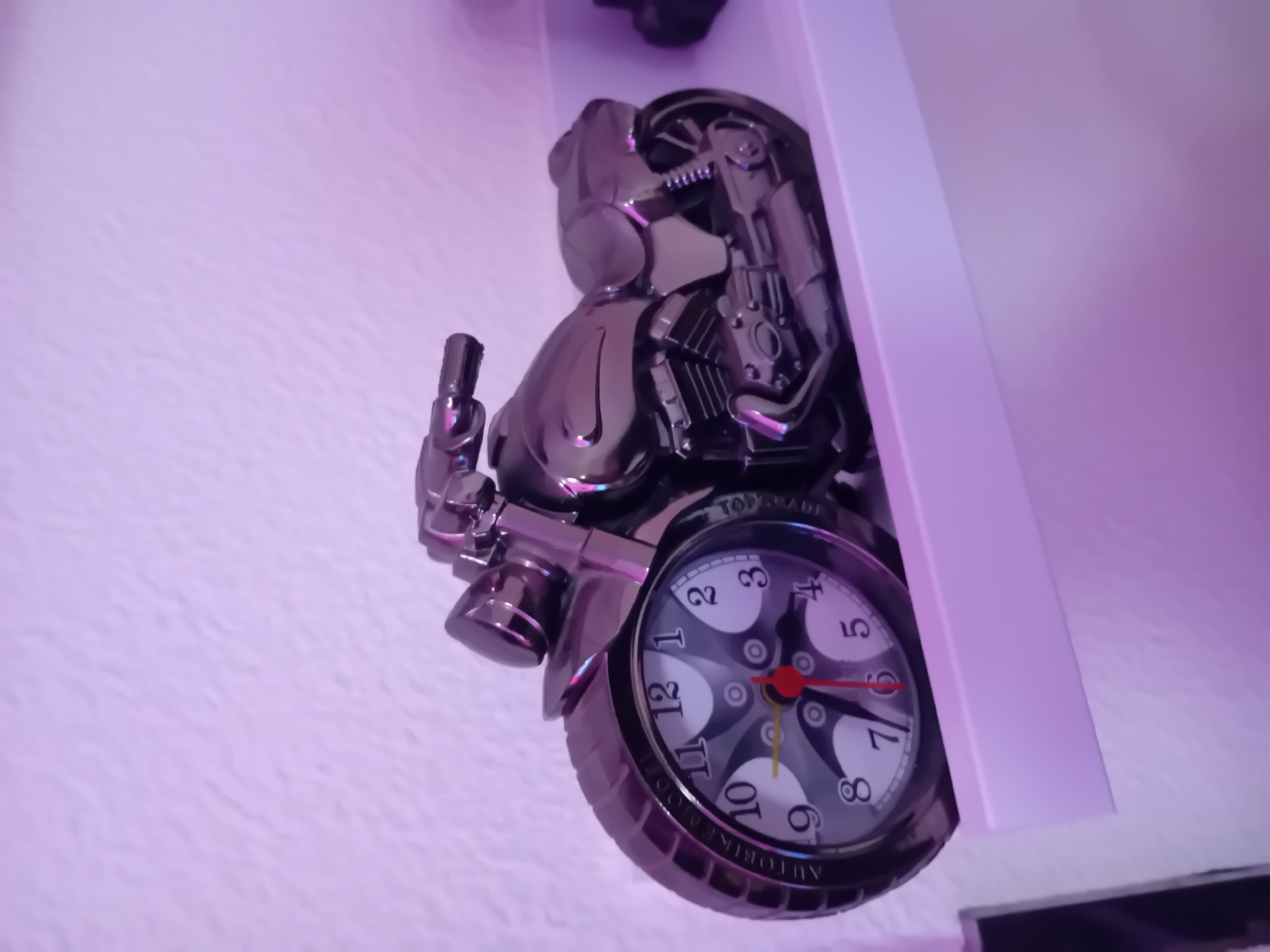 A low-angle shot depicts a motorcycle-themed wall clock mounted on a textured, off-white wall. The clock is mostly metallic and features a motorcycle design with the clock face integrated into the wheel. The face has black Arabic numerals and red and yellow hands. The base of the clock reads "AUTOBIRKE MODEL". The scene is illuminated with a pinkish-purple light.