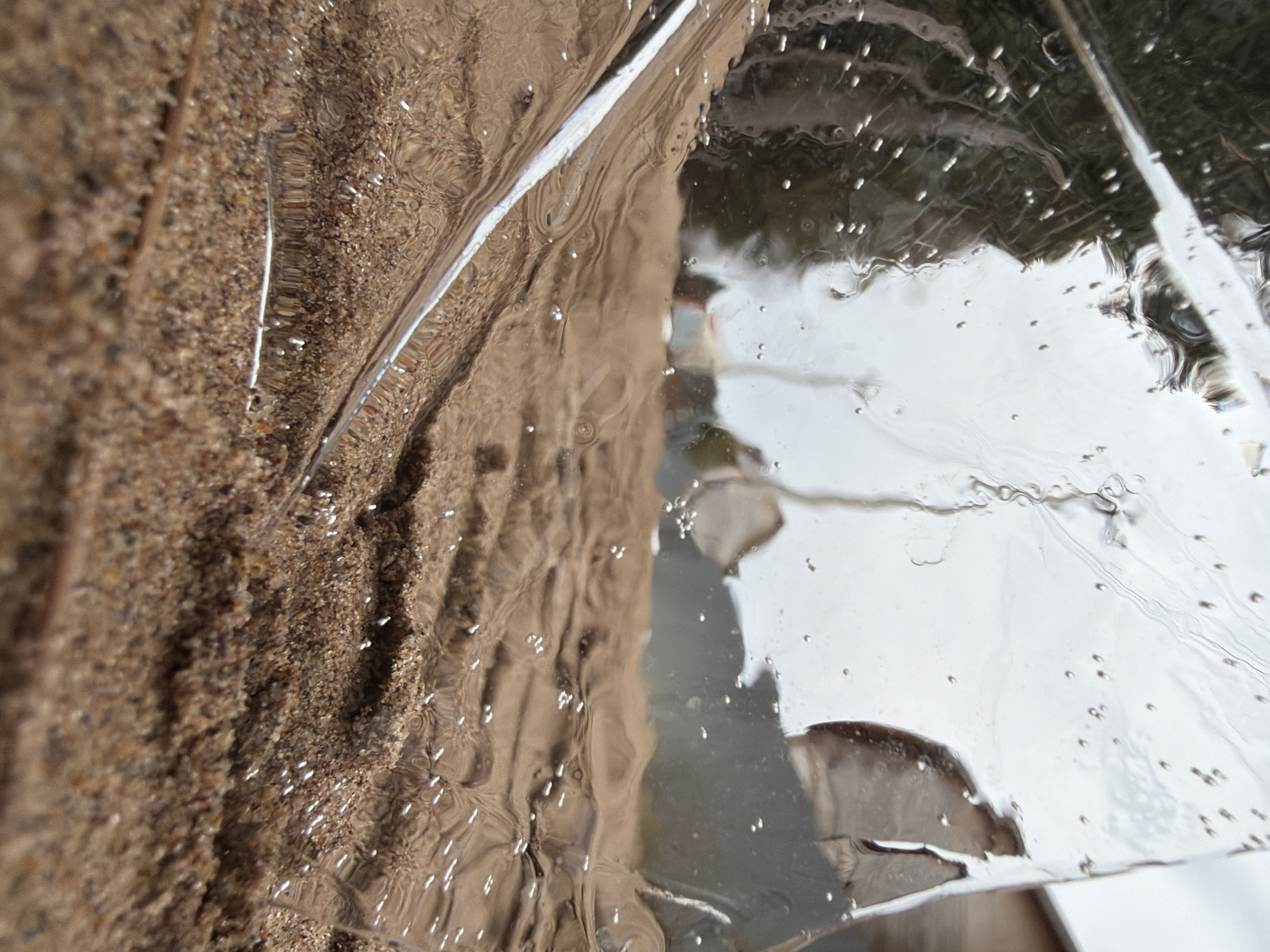The image shows a close-up view of shattered glass against a rough, sandy or earthen surface. The glass appears to have numerous small water droplets or imperfections on its surface, distorting the view of whatever lies beyond. Through the clear sections of the glass, hints of an outdoor environment are visible, possibly including vegetation and structures. The location is not discernible within the image.