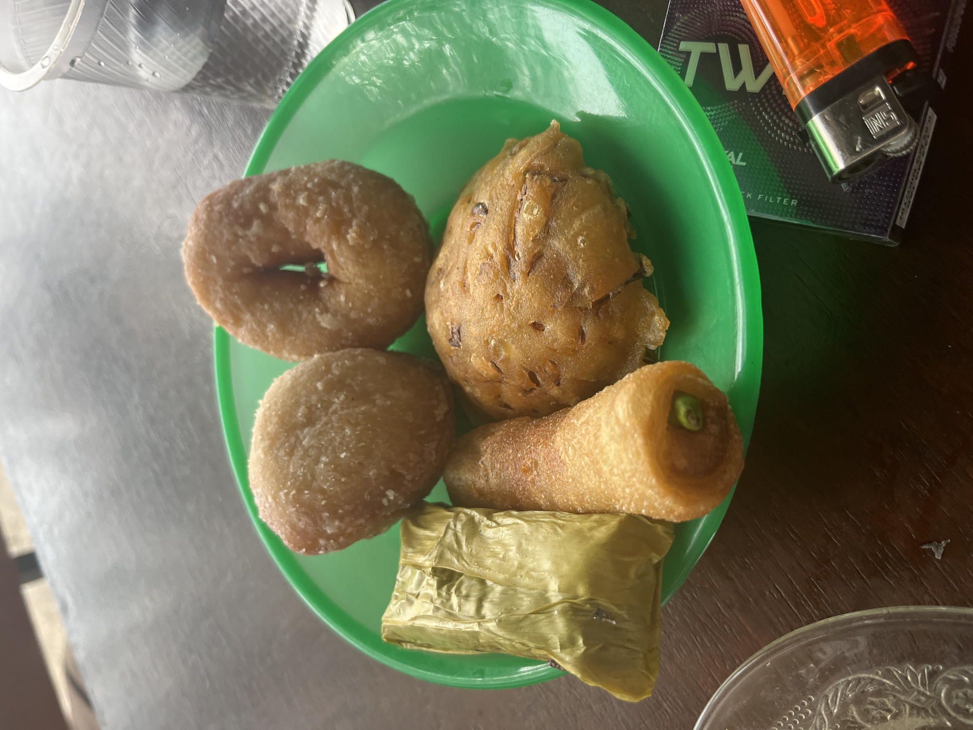 A plate of assorted Indonesian snacks sits on a wooden table. The snacks include two round, fried dough pastries coated in sugar, a golden-brown, cone-shaped fried item with a green pepper inside, a rectangular snack wrapped in a banana leaf, and a flattened, round fritter. To the right of the plate, a clear plastic cup contains water, and a pack of cigarettes with an orange lighter rests on the table. The scene appears to be outdoors in Keudematangkuli, Indonesia, indicated by the presence of tropical snacks and the general ambiance.