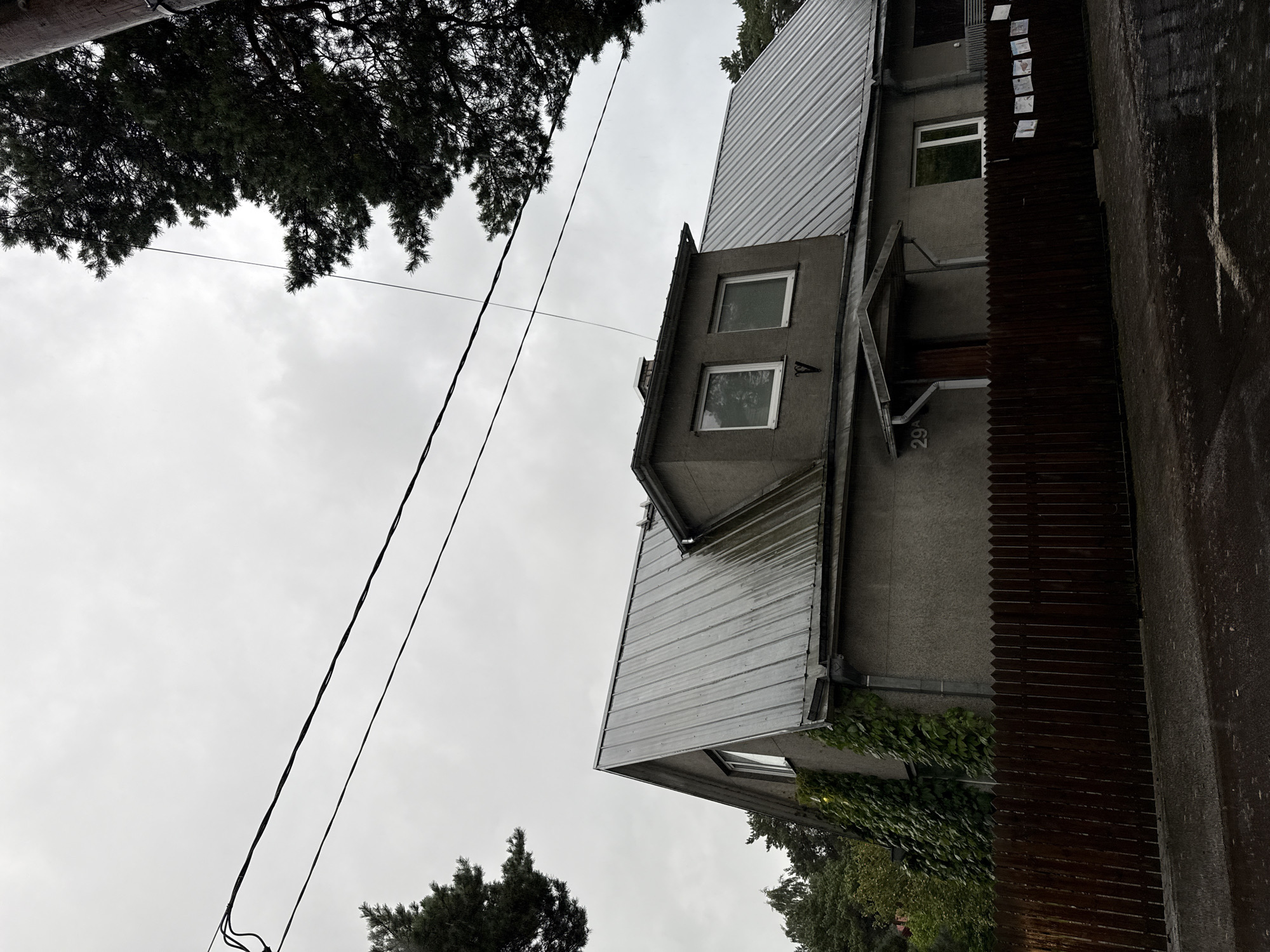 The image depicts a gray, two-story building with a metal roof, situated on a street in Tallinn, Estonia. The building has several windows, including two small square windows on an upper section and a larger rectangular window on the lower level. A wooden fence runs along the front of the property. There are overhead power lines visible in the sky, and trees are present on either side of the house. The ground appears wet, suggesting recent rain. There are no people visible in the image. The house is number 29A.