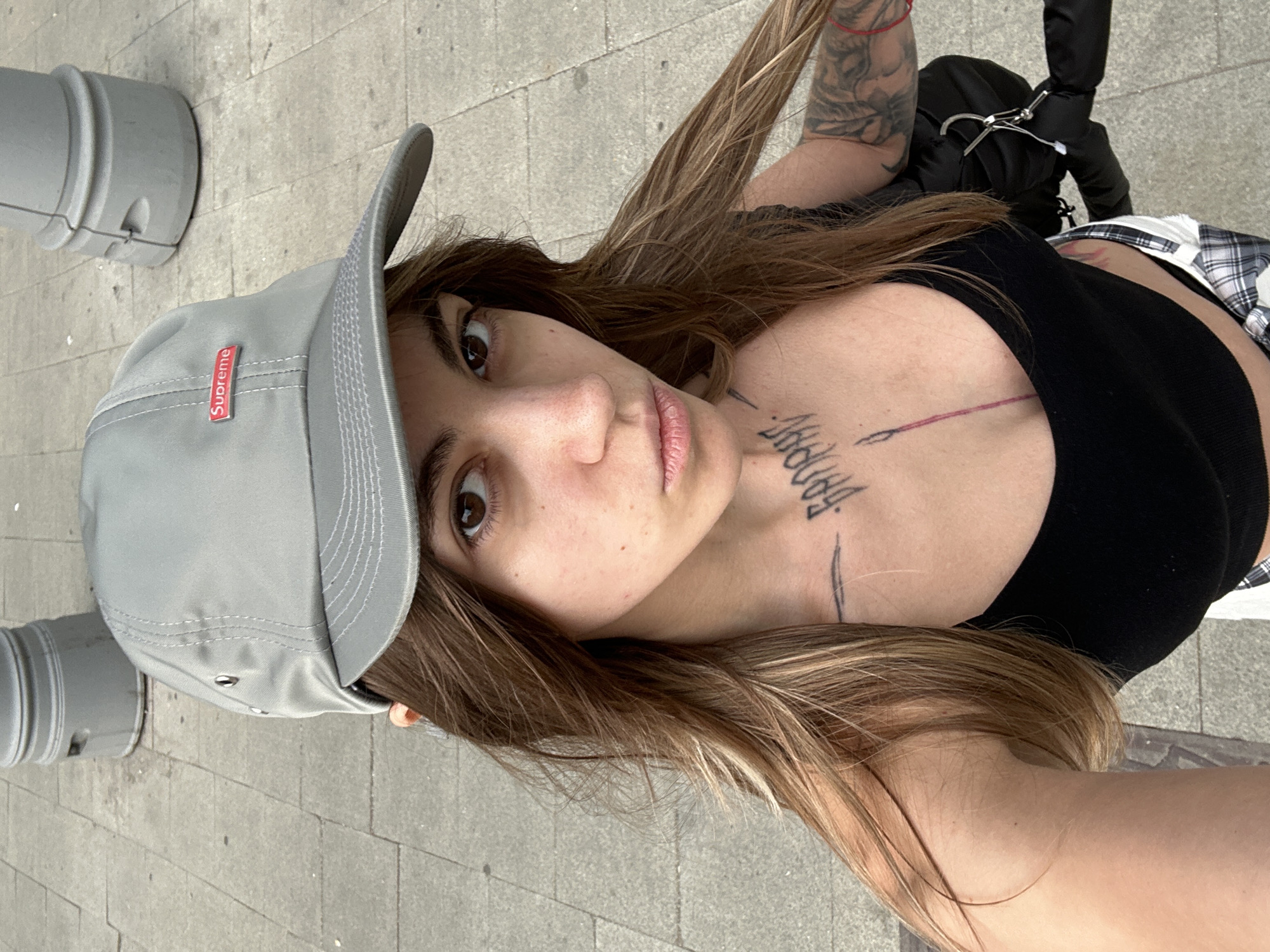 The image shows a close-up selfie of a young woman with long, brown hair, wearing a grey Supreme baseball cap and a black tank top. She has several tattoos, including a prominent one on her chest. She is standing outdoors on a paved surface, possibly a sidewalk or plaza, with a grey cylindrical object in the background. The lighting suggests it is daytime. The background details do not provide specific clues to indicate the location is Moscow, Russia.