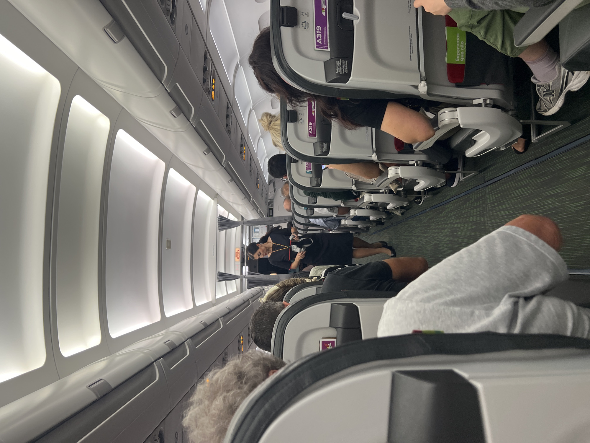 The image depicts the interior of an airplane cabin, viewed from the perspective of a passenger seated towards the rear. The cabin is illuminated by overhead lights set into the ceiling panels. Passengers are seated in rows on either side of a central aisle. The seats are upholstered in grey and white, with visible headrests. On the back of the seats, there are safety information cards and seat number identifiers like "A319".
A flight attendant, wearing a dark blue uniform dress and black shoes, is standing in the aisle and walking towards the front of the plane. She has her hair tied back and is holding what appears to be a tablet or a small device. She is looking forward as she walks. Some passengers are visible in their seats, some looking forward, others looking down, possibly at their phones or reading material. The floor of the aisle is carpeted in green.
The scene is consistent with a typical commercial flight. The presence of safety cards with specific seat numbers and the flight attendant in uniform indicate an operational aircraft during a flight or boarding process. There is no specific information within the image that would directly link it to the city of Moreira, Portugal.