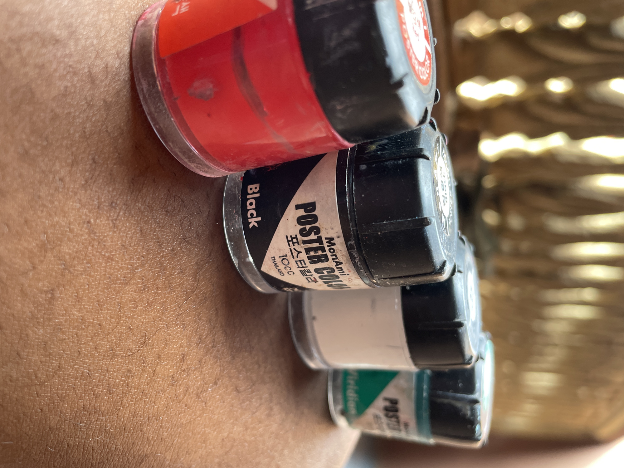The image shows a close-up of four stacked, small jars of paint. The topmost jar is red, followed by black, white, and green. The jars appear to be MonAmi Poster Color, as indicated by the labeling on the black jar. Text on the black jar also states "Thailand" and "10CC". The jars are resting on human skin. There is no evidence within the image to confirm or deny the location is Nkpor, Nigeria.