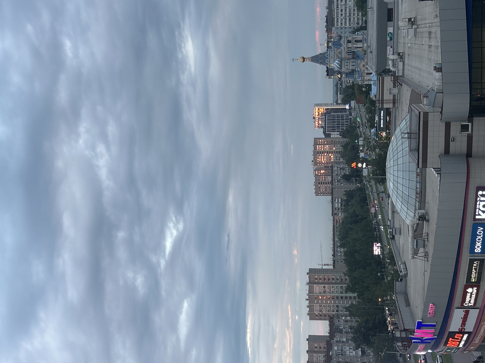 This is an aerial, exterior, full shot of a city scene in Izhevsk, Russia, under a cloudy, overcast sky with hints of pinkish-orange light reflecting off the clouds, suggesting either sunrise or sunset.
In the foreground, the roof of a large, multi-story building, likely a shopping center, is visible. It features a large, geodesic dome structure and various rooftop mechanical equipment. Signs are visible on the building's facade, including "KWT", "RBT.ru", "Семья", "Старик Хинкали", "НВАРТАЛ", and "SOKOLOV".
Beyond the foreground building, a city skyline is depicted. Several tall, apartment buildings with repetitive window patterns dominate the mid-ground, some of which are reflecting the warm sky light. Between the apartment buildings, a dense line of trees is visible. A road with vehicles is present in front of the apartment buildings.
To the right of the apartment buildings, a distinct blue and white church with golden domes stands out. This church is likely the Holy Trinity Cathedral, a prominent landmark in Izhevsk.
The overall impression is an urban environment at dusk or dawn, characterized by residential buildings, commercial structures, and a religious edifice under a dramatic sky.