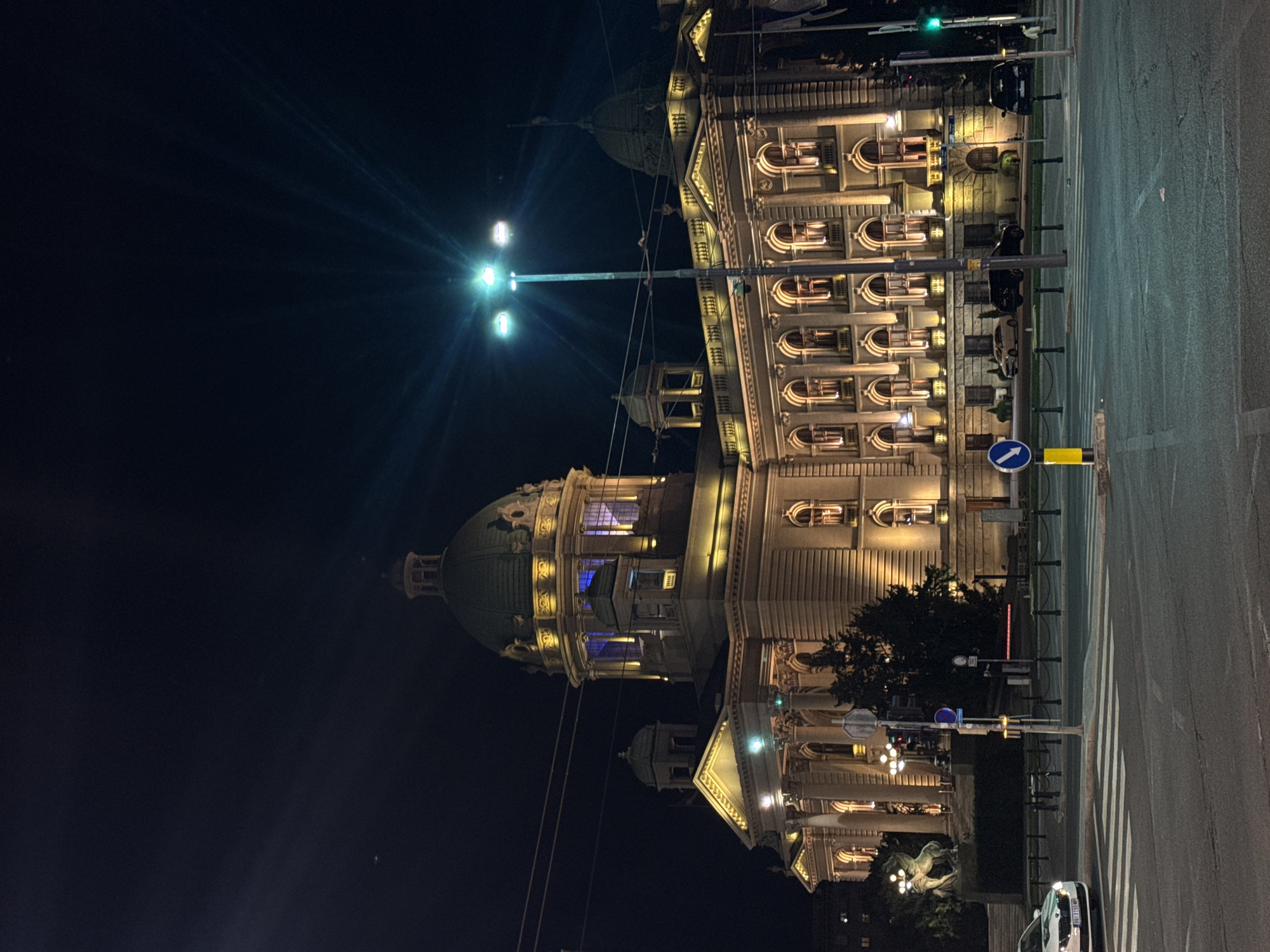 At night, the House of the National Assembly of Serbia is illuminated along a street in Vracar Urban Municipality, Serbia. The building is visible to its full extent, featuring its main dome, several smaller cupolas, and ornate window designs. Yellow light spills from the windows, accentuating the architectural details. A streetlamp casts a bright, star-like flare against the dark sky. A road runs alongside the building, with zebra markings for a pedestrian crossing. A sign with a blue arrow indicates direction. At street level, there are trees and shrubbery. A statue is illuminated by lamps. A white car is parked along the curb. A traffic light is green.