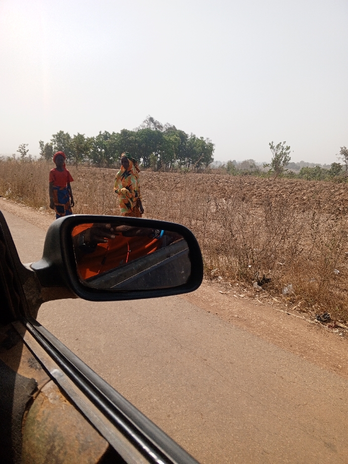 The image depicts a rural roadside scene, reportedly in Kuta, Nigeria, captured from the interior of a vehicle. In the foreground, the black plastic casing of a vehicle's driver-side mirror is visible, reflecting a person wearing an orange garment, potentially holding a mobile device. Below the mirror, a portion of the vehicle's window frame and a weathered metal body panel are seen.
On the right side of the image, an asphalt road runs parallel to the vehicle. Adjacent to the road is a dirt shoulder, transitioning into a field of disturbed earth with dry, sparse vegetation. Two individuals stand on this shoulder. The person on the left is a smaller individual, possibly a child, wearing a red short-sleeved top, a blue and white patterned wrap skirt, and a red head covering. The person on the right is an adult female, dressed in a vibrant multi-colored patterned dress or wrap (featuring yellow, green, orange, and blue hues) and a matching head covering. She holds an indistinct object, possibly a bundle or bag, in her left hand. Both individuals are facing generally towards the road and the vehicle's direction of travel.
The background features a line of green trees bordering the field, with a hazy, light-colored sky above. The overall environment suggests a dry, possibly agricultural, rural landscape. No explicit interactions between the individuals or the vehicle are discernible.