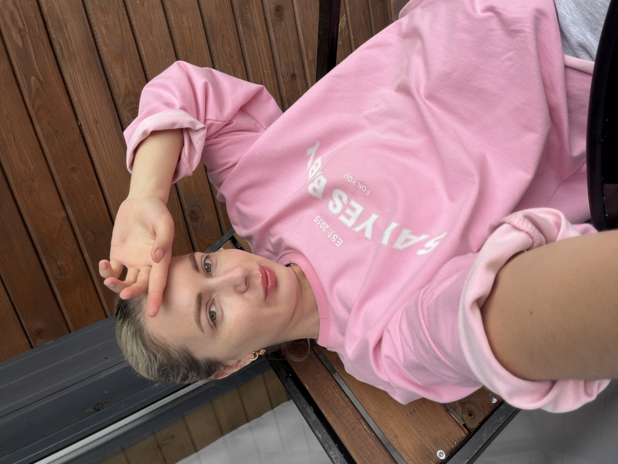 A Caucasian woman with blonde hair pulled back is seated on a wooden bench outdoors. She is wearing a pink sweatshirt with white lettering that reads "EST. 2015 SAY YES BABY FOR YOU". Her right hand is raised to her forehead. The background consists of wooden siding and a dark gray structure. The image is taken from a slightly high angle.