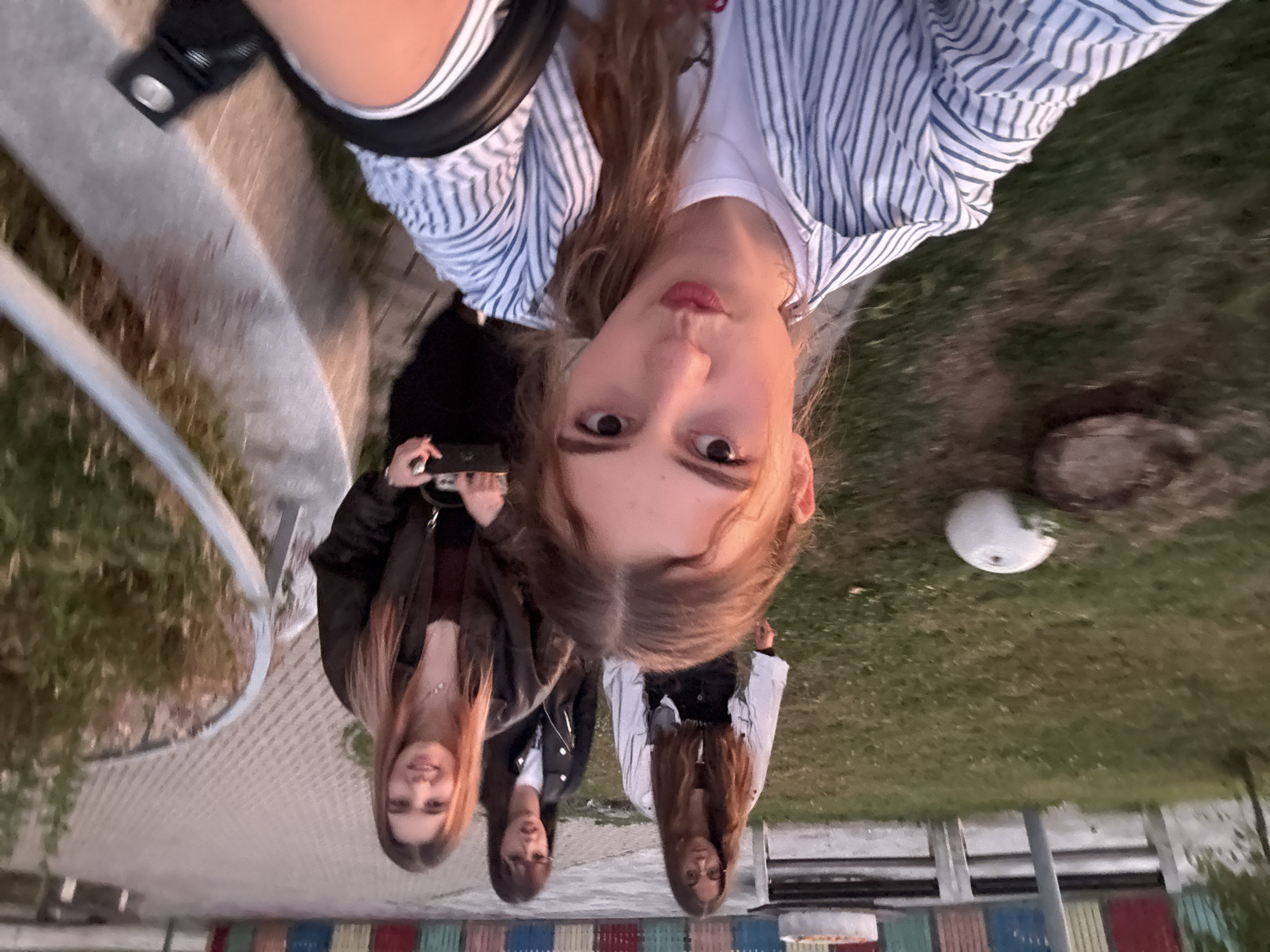 Four young women are gathered outdoors in what appears to be a park or public area, likely in Panagyurishte, Bulgaria, given the context. The woman in the foreground is facing the camera and taking a selfie, her face is close to the lens and she is making a puckered expression. She is wearing a white and blue striped button-down shirt over a white t-shirt. Behind her, three other women are visible. One woman in a black leather jacket and white t-shirt is looking towards the camera, while another woman in a light-colored jacket with a black inner garment stands to her left, also looking towards the camera. The fourth woman is further back and slightly out of focus. The background shows a grassy area, a paved walkway with a railing, and colorful structures that could be part of a playground or decorative installation. The lighting suggests it is late afternoon or early evening.