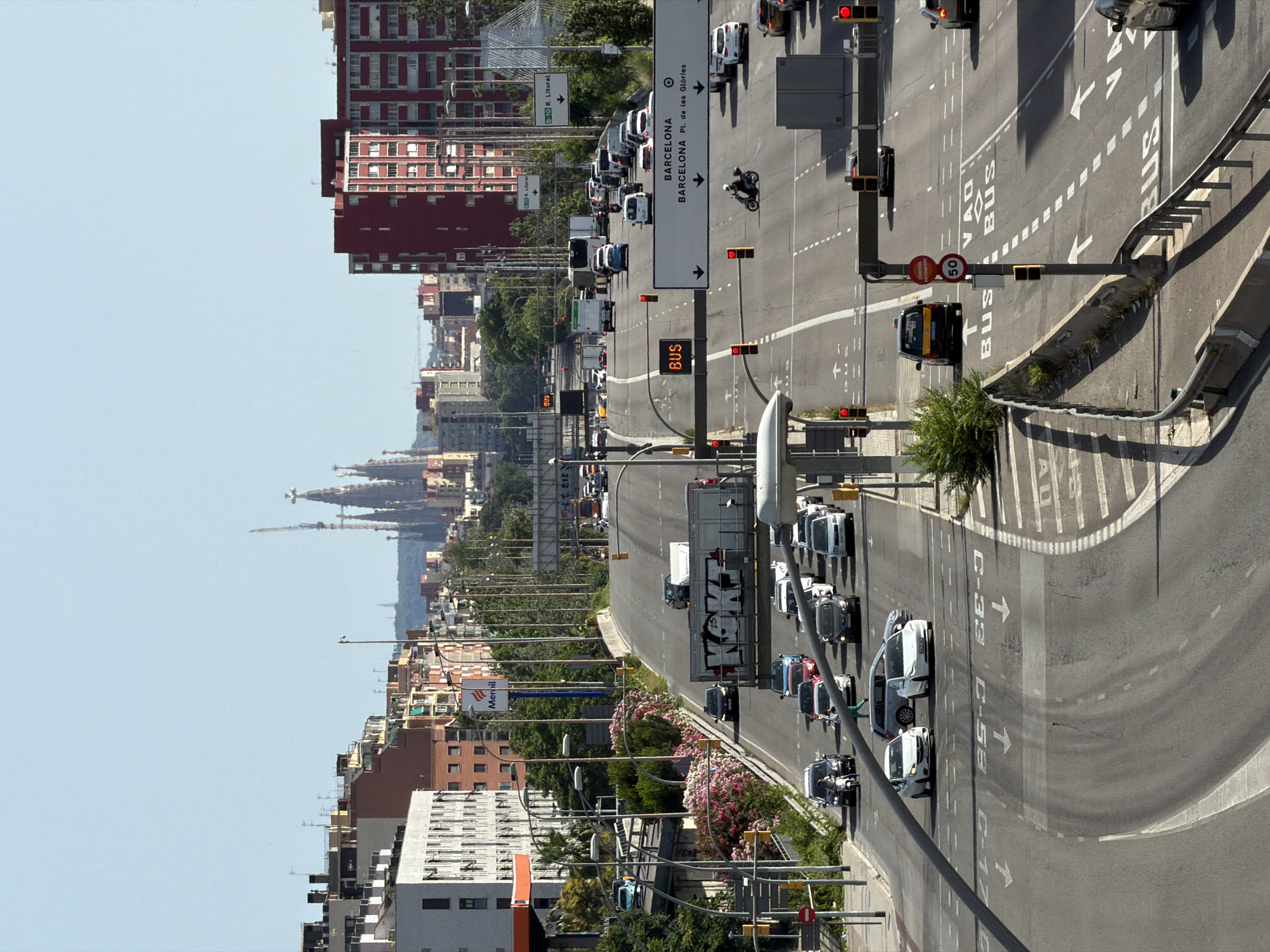 The image captures an aerial view of a multi-lane highway in Barcelona, Spain, on a clear, sunny day. The highway is bustling with traffic, including cars, trucks, and a motorcycle. In the foreground, several lanes of traffic are visible, with cars moving in both directions. Road markings, including lane lines, arrows, and signs indicating "BUS," are clearly discernible. Traffic lights are present and appear to be functional.
A notable landmark in the background is the Sagrada Familia basilica, its distinctive spires visible against the sky, indicating the general location within the city. Various buildings, including residential apartments and commercial structures, line the sides of the highway, with the red-colored building being particularly prominent. Greenery, including trees and bushes, also features in the scene, particularly along the edges of the highway. A Meroli gas station sign is also visible on the left side of the highway. The overall impression is that of a busy urban environment with ongoing traffic flow.