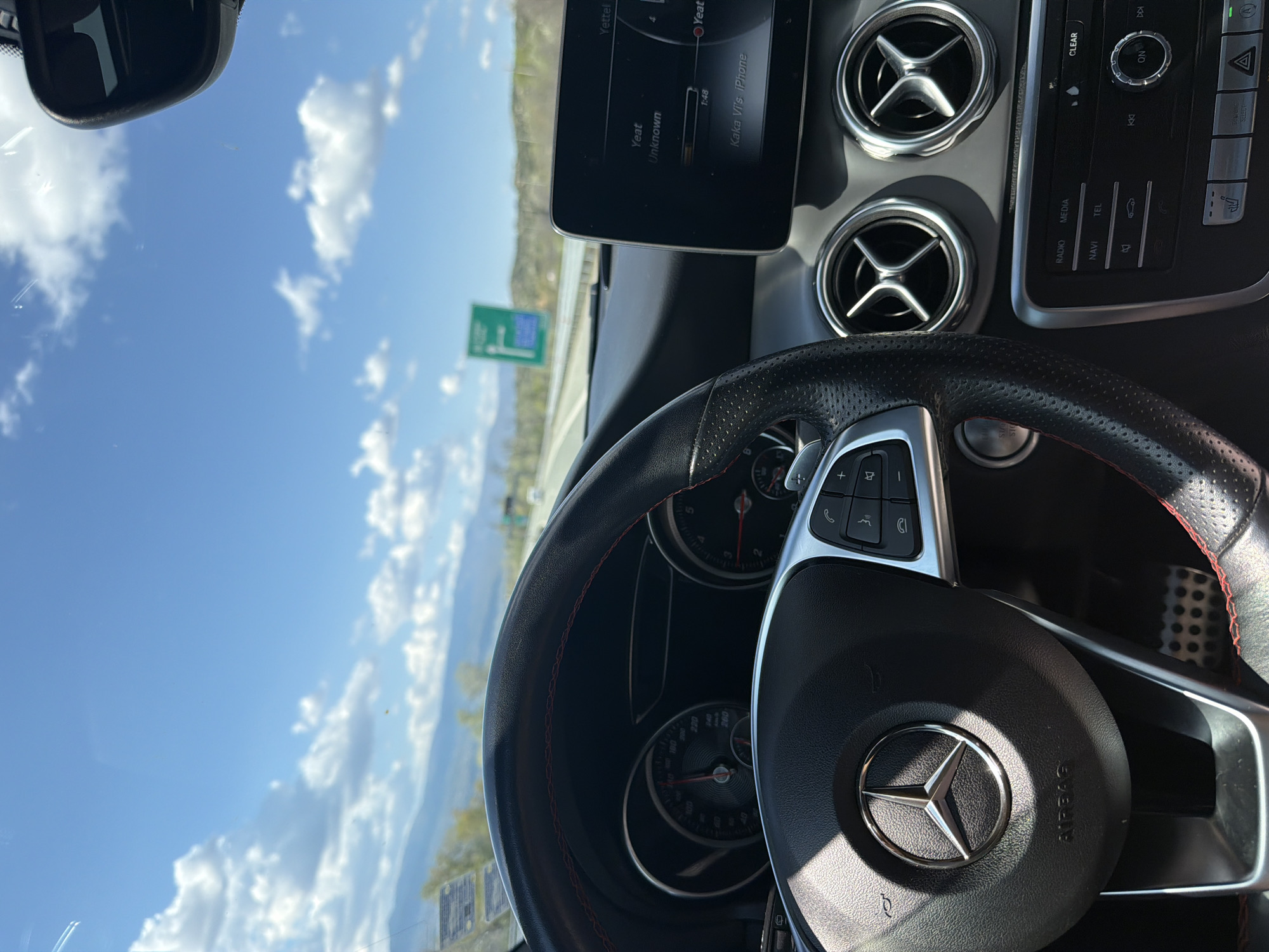 The image shows the interior of a Mercedes-Benz car, focusing on the dashboard, steering wheel, and a view through the windshield. The sky is blue with some clouds. The car's infotainment screen displays music information, and the dashboard features air vents. A road sign is visible through the windshield. The location in Valkovo, Bulgaria is not discernible from this image.