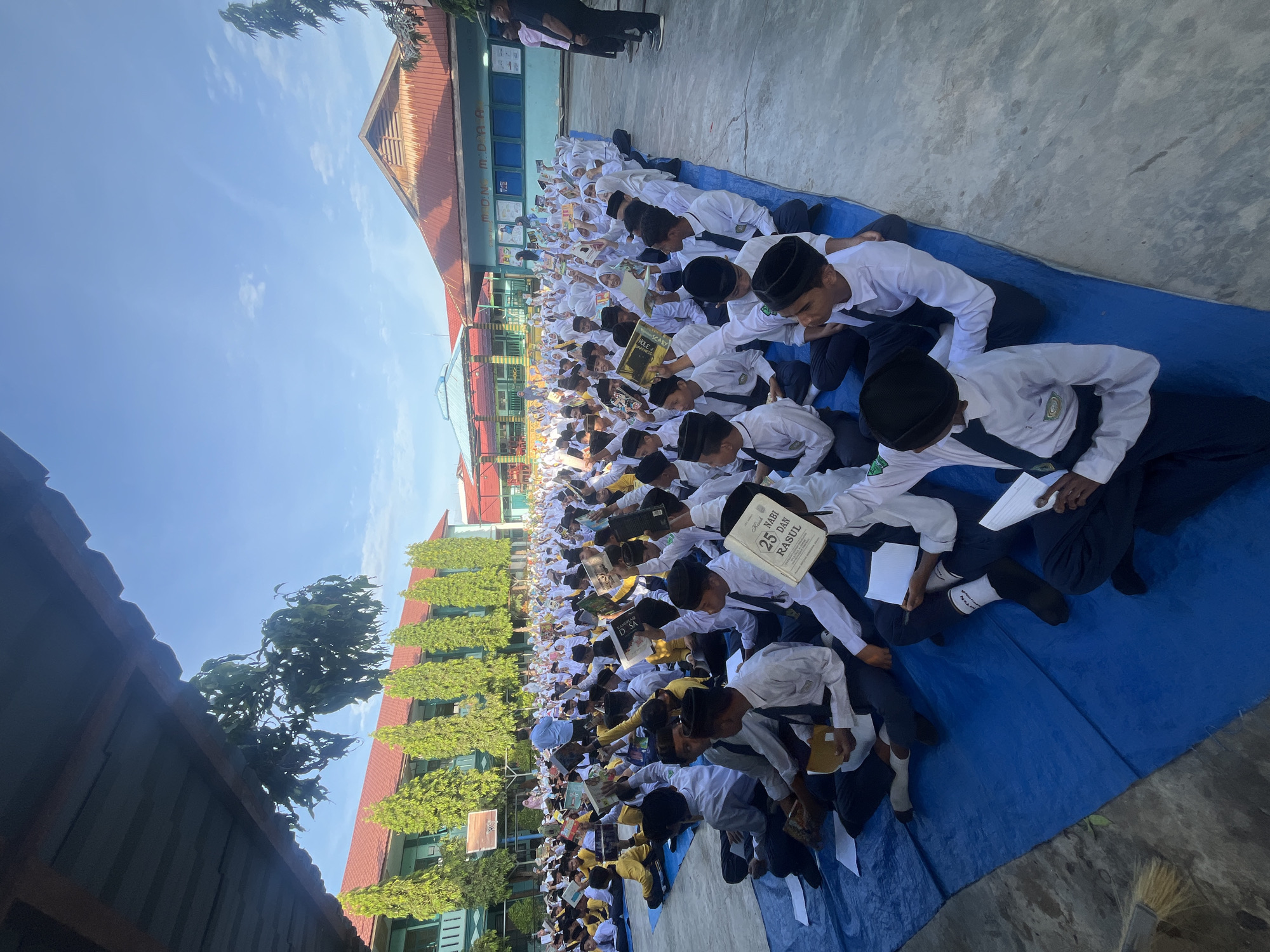 In Lhokseumawe, Indonesia, a large group of students, predominantly male and dressed in white shirts, black pants, and black caps, are seated on blue tarps outdoors. They appear to be participating in an educational activity, with many holding books or papers. The students are arranged in rows on a paved courtyard, facing forward towards an unseen focal point. Behind them, a building with green walls and red roofs is visible, possibly a school. The sky is clear and blue, with a few scattered clouds. The scene suggests a gathering for a lesson, assembly, or ceremony.