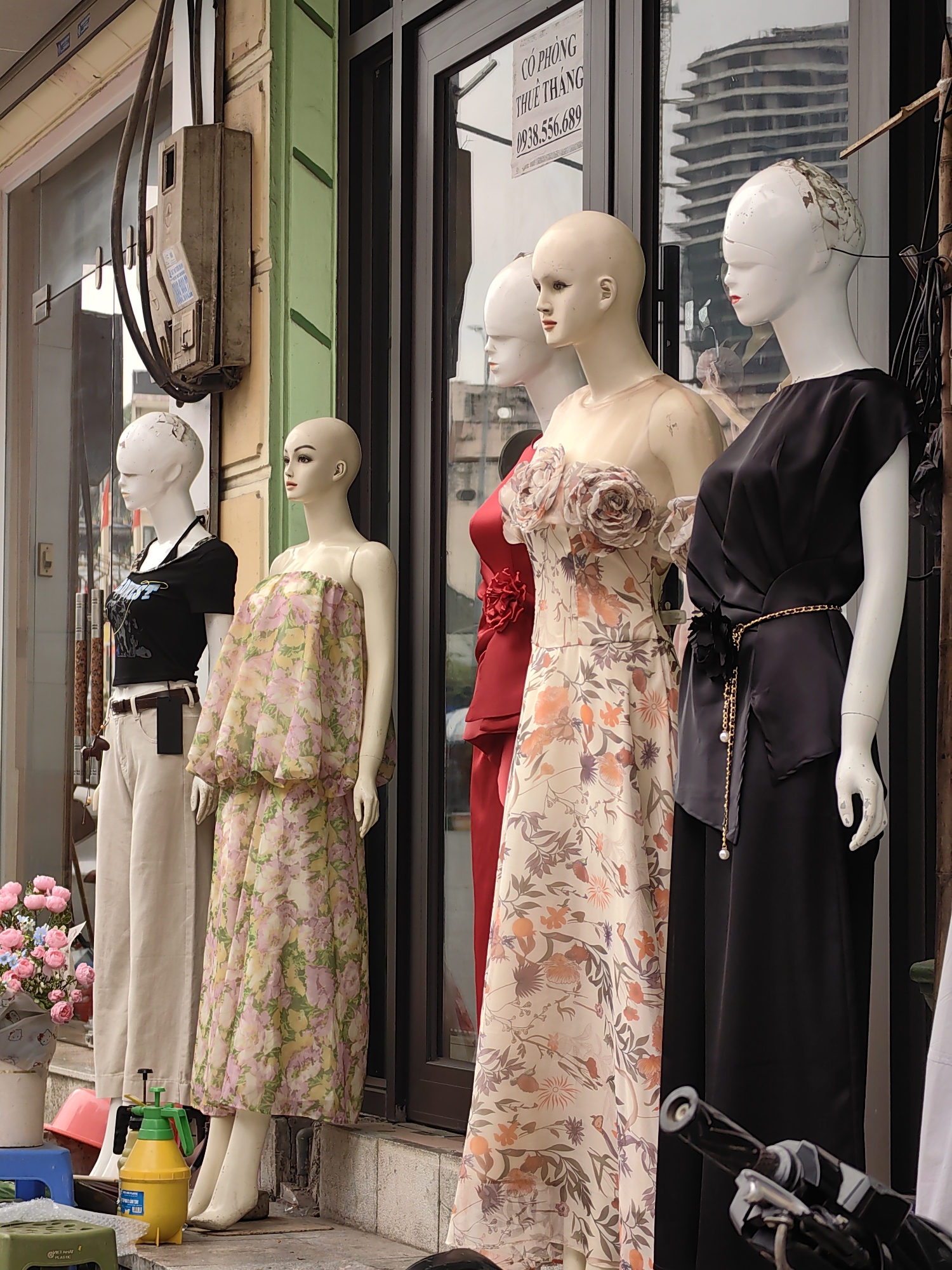 The image depicts a storefront in Bai Chay Ward, Vietnam, featuring five mannequins displayed outside. They are positioned in front of a building with green accents and a glass facade. The mannequins are adorned with different clothing items: one wears a black top with tan pants, another a floral print dress, the next a red dress, followed by a floral dress with decorative floral embellishments, and the last a black dress with a gold chain belt. A sign with Vietnamese text and a phone number is visible on the glass. Miscellaneous objects, including a yellow plastic sprayer and blue stools, are scattered on the sidewalk. The reflection in the glass shows a modern multi-story building under construction.