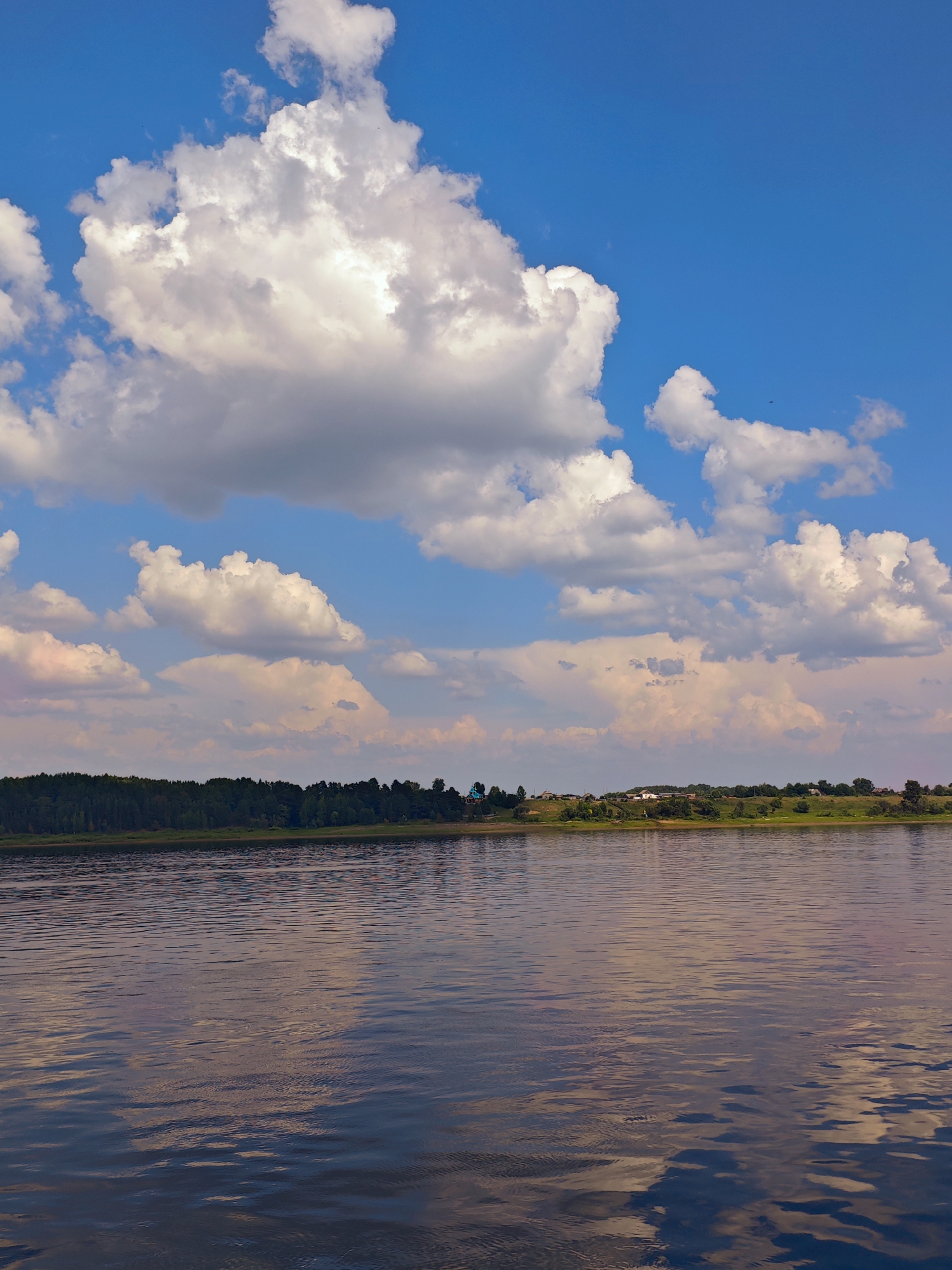 This is a vertical, eye-level shot of a wide, calm river reflecting the sky and the distant tree line and village. The water's surface is rippled, creating streaks of light and shadow. The sky is a bright blue with large, fluffy white clouds scattered across it, some tinged with pink from the setting sun. A dense line of dark green trees borders the far bank of the river, and behind them, a small village with scattered houses and rooftops is visible, nestled on a gently rolling green hill. The overall impression is one of peaceful rural scenery. The location is Erkalovo, Russia.