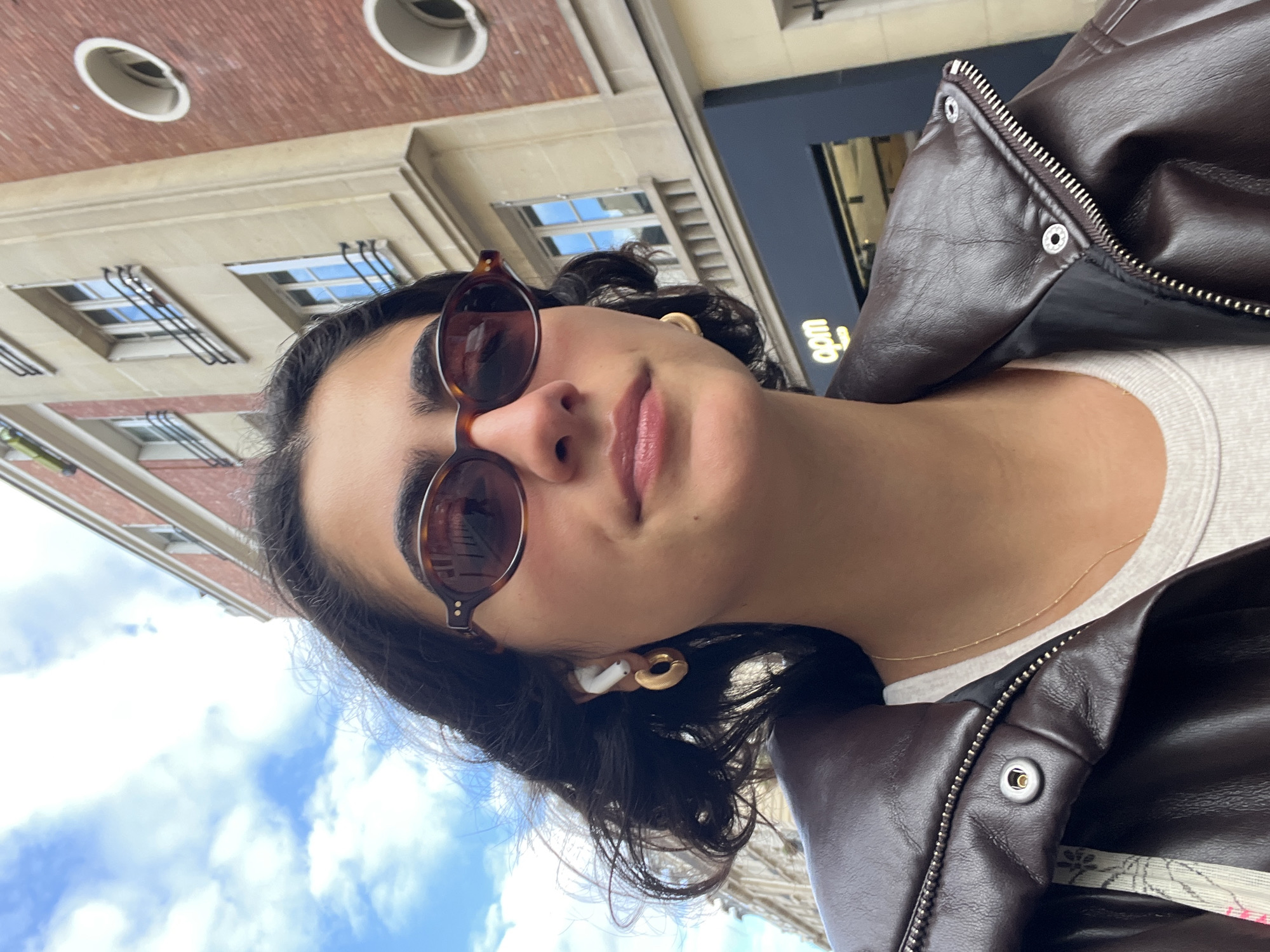 A young person is taking a selfie in Paris, France, on a partly cloudy day. The subject is wearing a dark brown leather jacket over a light gray shirt, with round brown sunglasses. An AirPod is visible in their ear, along with a gold hoop earring. In the background are Parisian buildings with distinctive architectural details such as circular ventilation openings and rows of windows with grilles. The sky is visible between the buildings, showing blue patches and white clouds. The person is looking slightly upward at the camera.