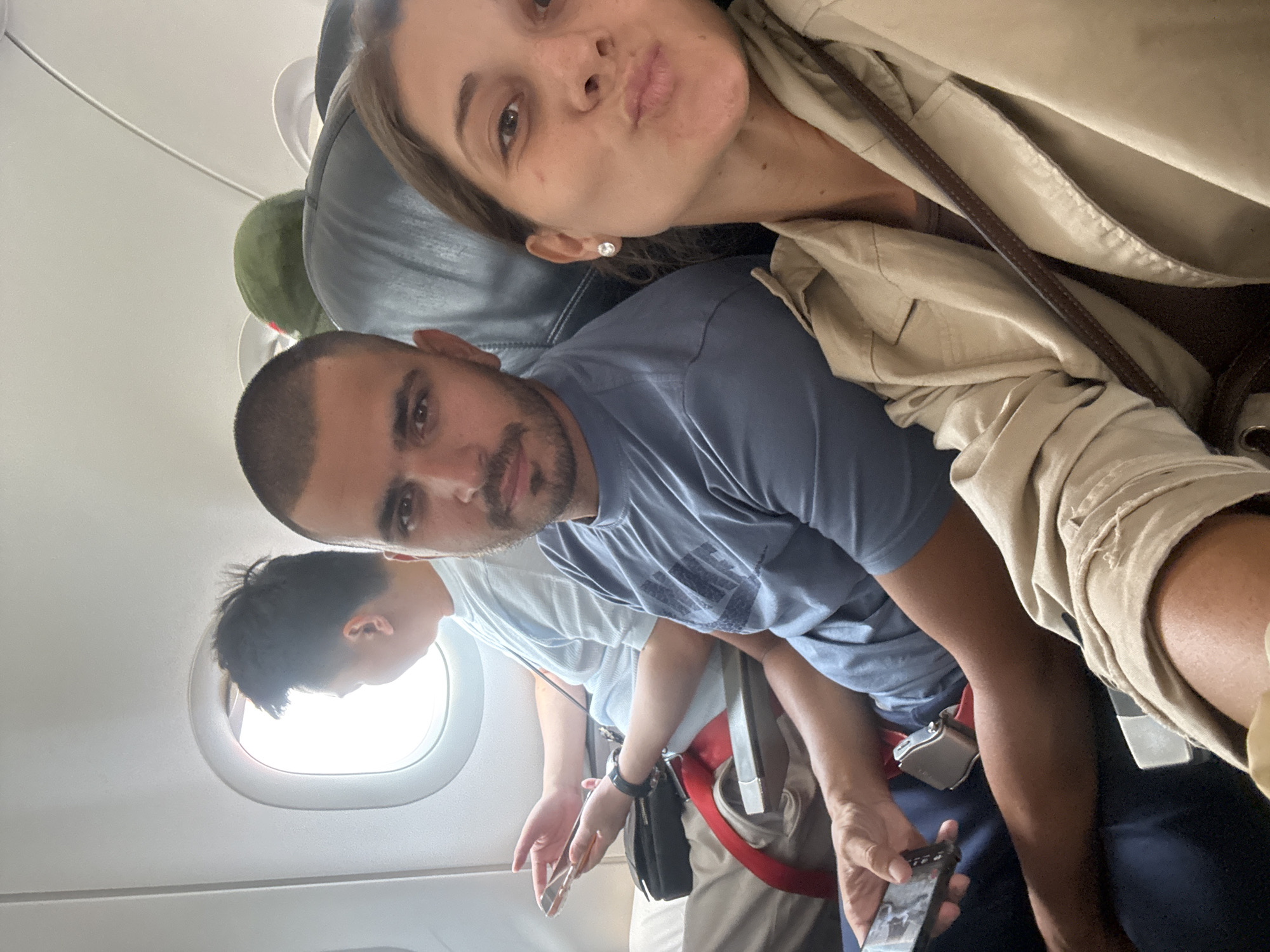 The image is taken from inside an airplane, looking towards the front. A woman with light brown hair and a tan jacket is in the top of the frame, puckering her lips for a selfie. Below her, a man with dark hair and a beard wears a blue shirt. In the background, another person looks out the window. A red seatbelt strap is visible. The location is not discernible from the image.