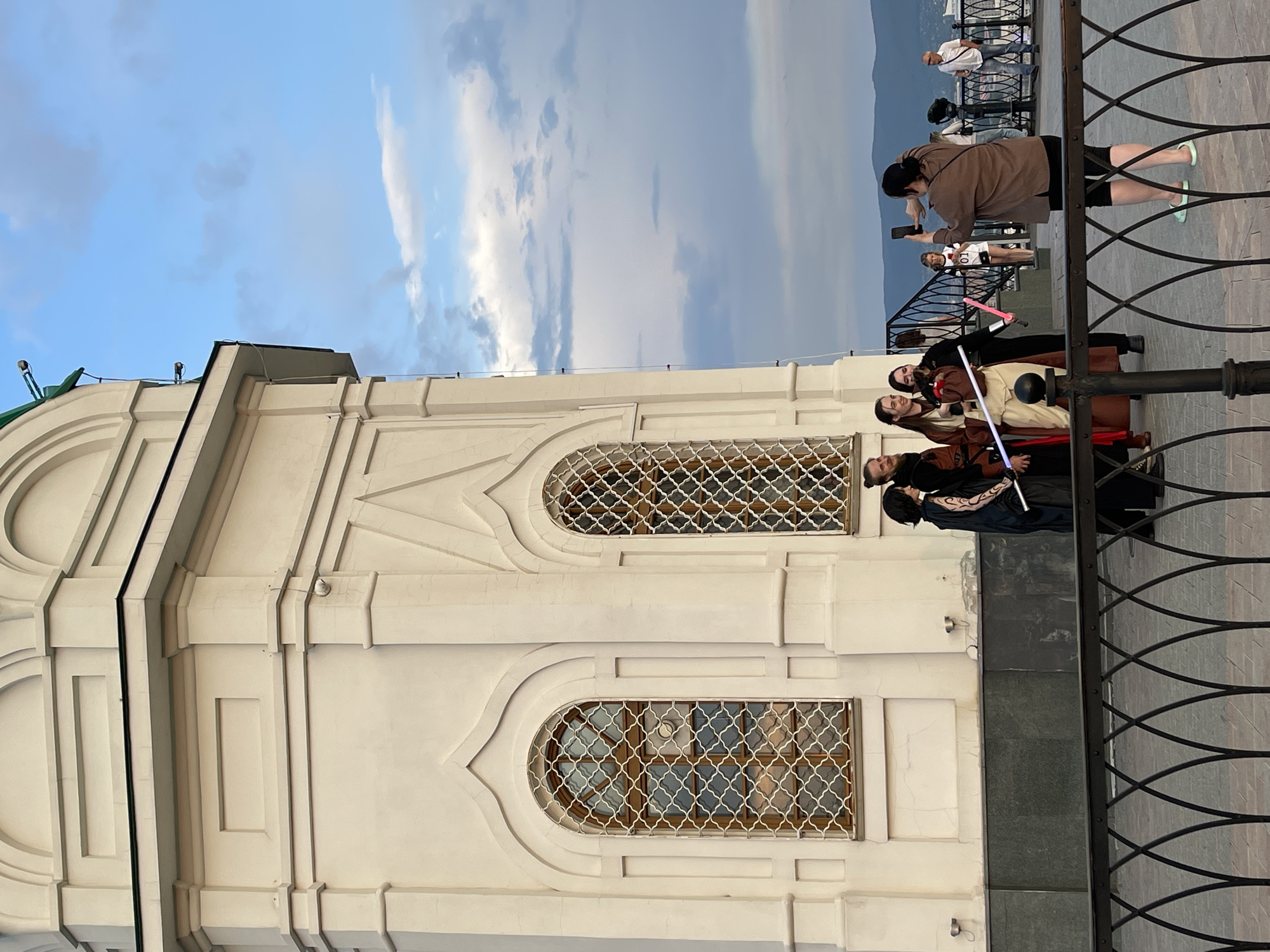 The image depicts a group of people in costumes, some holding illuminated sabers, posing in front of a white building. The building appears to be a historical or religious structure with arched windows and decorative architectural elements. In the background, there is a city skyline with mountains and a cloudy sky.
The location is the Paraskeva Pyatnitsa Chapel in Krasnoyarsk, Russia. The chapel, built in 1805, is a prominent landmark situated on a hill overlooking the Yenisei River and the city. It is a popular tourist destination and a symbol of Krasnoyarsk.
In the foreground, a woman is taking a photograph of the costumed group with her smartphone. The costumed individuals are likely participating in a cosplay event or a themed gathering. The sabers suggest a Star Wars theme.
The environment is outdoors, with a paved walkway and a decorative railing in the foreground. The weather appears to be overcast with some patches of blue sky. The overall atmosphere suggests a casual and festive gathering.