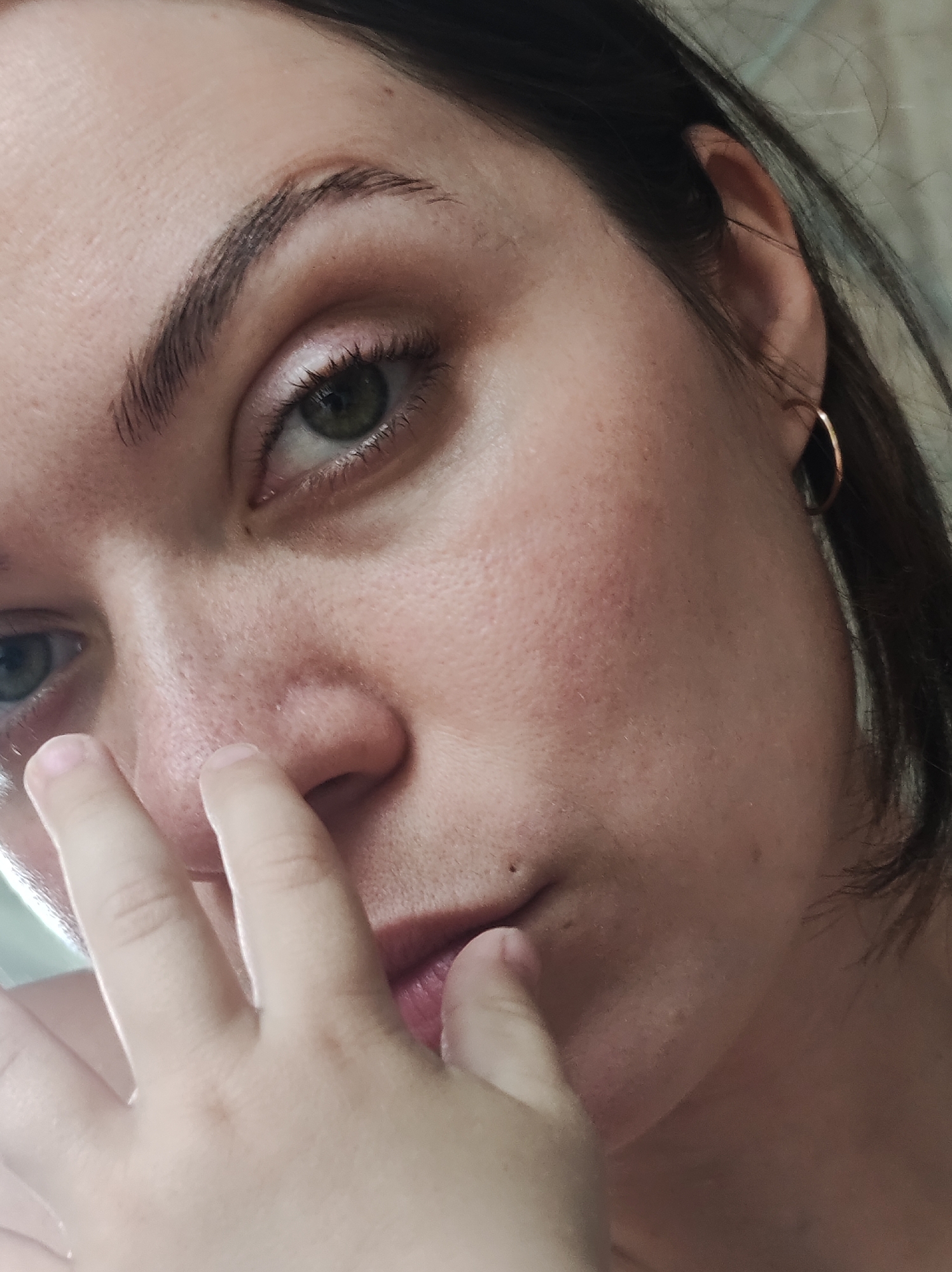 A close-up, slightly angled, selfie-style image of a woman's face. She has fair skin, dark brown hair, and green eyes. The woman is wearing small gold hoop earrings. Her right hand is partially obscuring her mouth. The background is out of focus. There is no definitive information in the image to suggest a location in Фоки, Russia.