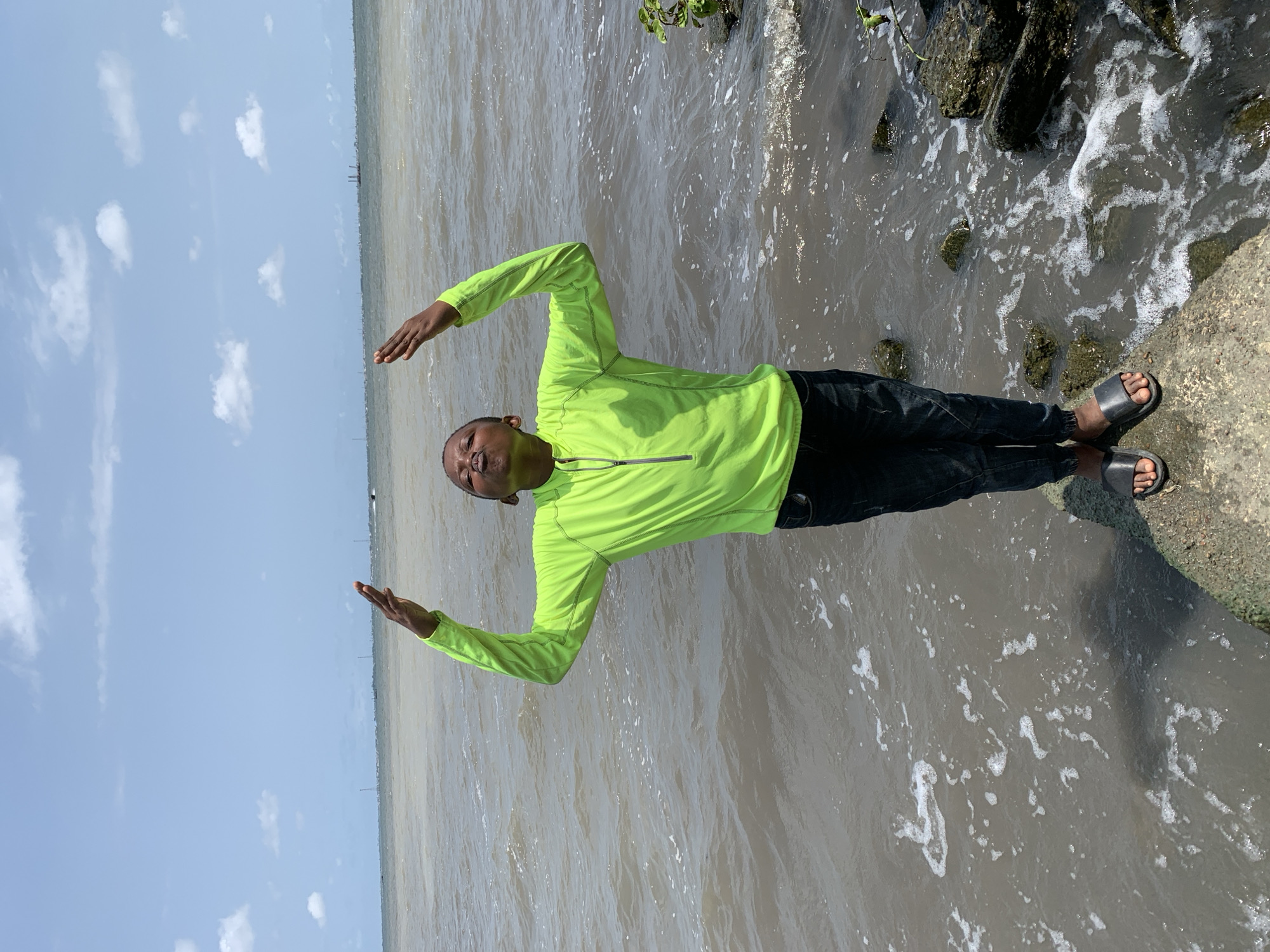 A man is standing on a concrete structure along a body of water. He is wearing a bright green long-sleeved shirt, dark pants, and sandals. His arms are outstretched, with his hands raised. The water appears murky and has some foam. In the distance, the sky is visible with a few clouds. The image appears to be taken on a sunny day. The location is likely near a shoreline in Ikorodu, Nigeria.