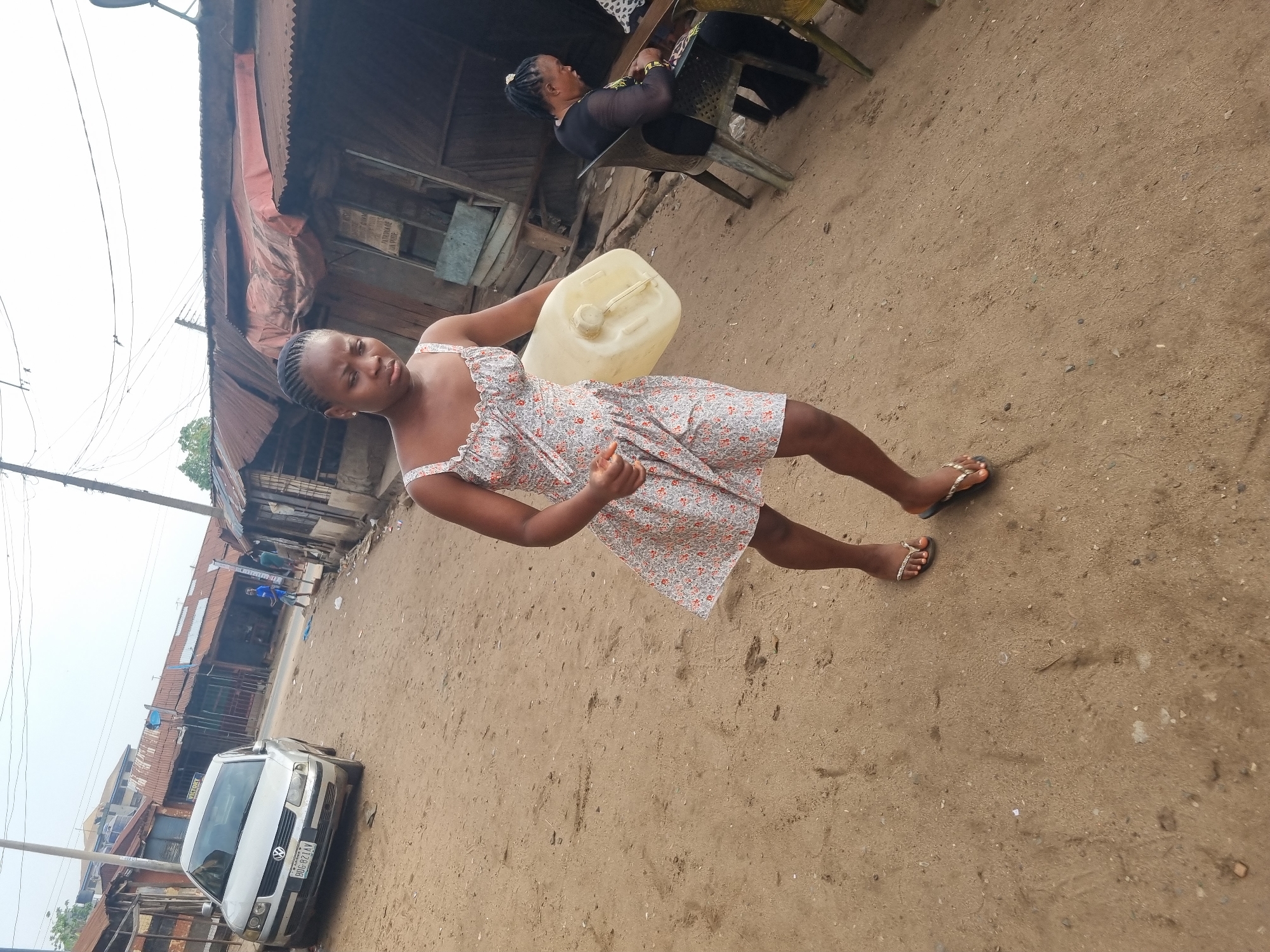 This image depicts a bustling street scene in Onitsha, Nigeria, under a bright daytime sky. The primary subject is a young woman, likely in her late teens or early twenties, standing on an unpaved, dusty road. She looks directly at the camera with a composed, neutral expression. Her dark hair is styled in braids, and she wears a sleeveless floral-patterned dress and flip-flops. She holds a yellow plastic jerrycan in her left arm, suggesting daily activities like water collection or transport.
In the background, a second woman is seated on a plastic chair outside a building, looking towards the left. She wears a dark, long-sleeved top. The street is lined with various buildings. On the left, simpler structures made of wood and corrugated iron, many with rusted roofs, indicate a common architectural style in the region. Utility poles with numerous overhead wires crisscross the sky.
On the right side of the road, a white Volkswagen Passat car is parked, with its license plate "BDG-827AY" clearly visible, along with the word "LAGOS" below it. Beyond the car, more buildings with roll-up metal doors and corrugated roofs line the street, likely serving as small shops or residences. The overall scene portrays an everyday, active environment in an urban or semi-urban area of Nigeria, characterized by local architecture and community life. The dry, dusty ground reinforces the outdoor setting.