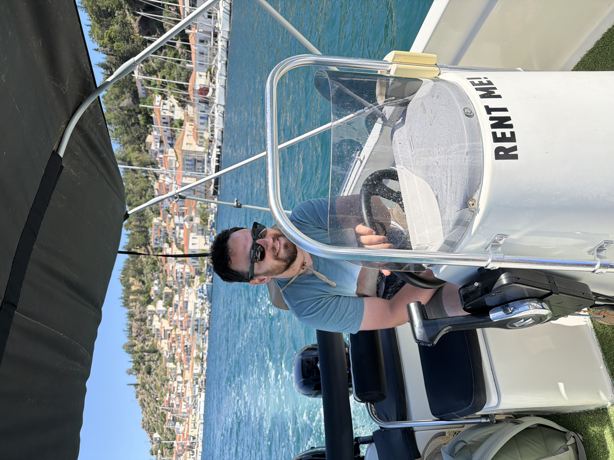 A man is depicted driving a small motorboat on a sunny day, with the scenic town of Poros, Greece, visible in the background. The man, with short dark hair, a beard, and sunglasses, is seated at the helm, holding the steering wheel with his right hand. He is wearing a light blue t-shirt and a beige sun hat is tied around his neck. A wide smile suggests he is enjoying the experience.
The boat is a white rental vessel, indicated by the black text "RENT ME!" prominently displayed on its console, located to the right of the steering wheel. The throttle control is visible to his left. A dark green or black canvas canopy partially covers the boat, providing shade. Below the console, a grey backpack or bag is visible on the boat's floor.
The water in the foreground and midground is a clear, bright turquoise, showing ripples from the boat's movement. In the background, a picturesque coastal town built into a green, tree-covered hillside stretches across the horizon. The buildings feature traditional Mediterranean architecture, with light-colored walls and terracotta roofs, characteristic of Greek islands. The sky above is a clear, vibrant blue, indicating fair weather. The overall scene conveys a cheerful and leisurely summer day spent boating in a beautiful Greek setting.