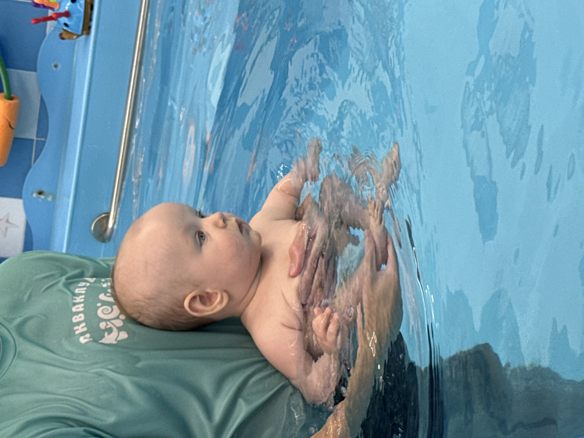 An overhead shot shows a baby in a swimming pool, held by an adult. The baby is bald with blue eyes and light skin. The adult, wearing a teal t-shirt with white lettering that appears to say "AKBAKU," is supporting the baby under its arms. Both are submerged in clear blue water. The pool has a blue tiled edge, and in the background, a metal rail is visible, along with children's toys including an orange float and a blue toy with red antennae. The image is likely taken at a baby swimming class in Moscow, Russia, given the text on the t-shirt.