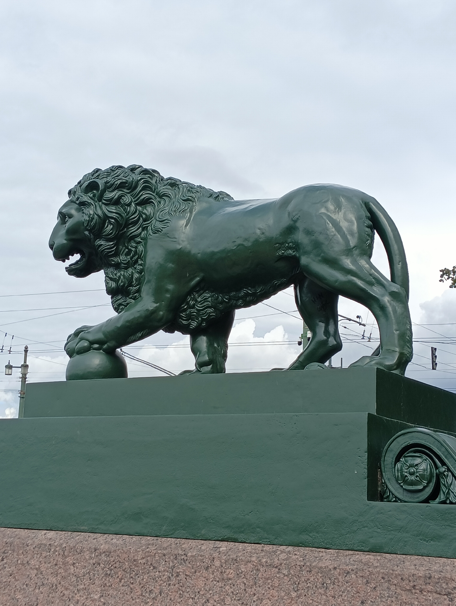 A dark green, metallic sculpture of a lion is positioned on a matching green stone pedestal. The lion is depicted in a powerful stance, with its mouth open and its front left paw resting on a sphere. The sculpture is situated outdoors, with a cloudy sky in the background. Overhead wires and a lamppost are visible in the mid-ground, suggesting an urban setting. The pedestal appears to be part of a larger structure, possibly a bridge, given the architectural flourish on the right side of the image. This sculpture is located on the Lions Bridge in St. Petersburg, Russia, known for its four lion sculptures that guard the entrance to the Kryukov Canal.