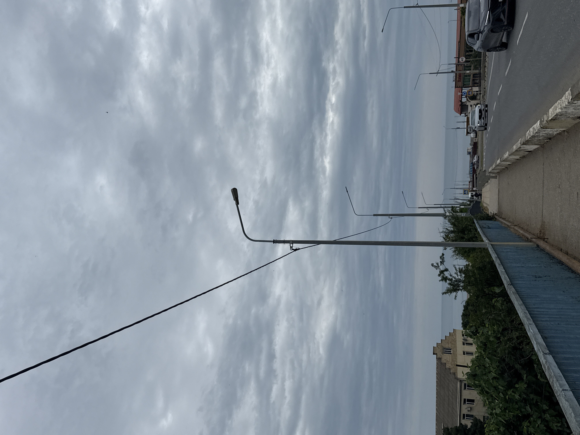 This media file depicts a cloudy day in Derbent, Russia. The scene is an outdoor urban environment with a street, sidewalk, and a body of water in the distance. A power line runs diagonally across the frame. Several streetlights are visible, suggesting it is either dawn, dusk, or an overcast day. Cars are present on the road, indicating typical urban activity. The architecture of a building in the background appears to be consistent with Russian style. The overall mood is neutral, dominated by the overcast sky and mundane street elements. There are no people prominently visible, and the focus is on the urban landscape and the atmospheric conditions. The time of day is not clearly defined but is likely during daylight hours due to the activity on the street. The weather is clearly cloudy.