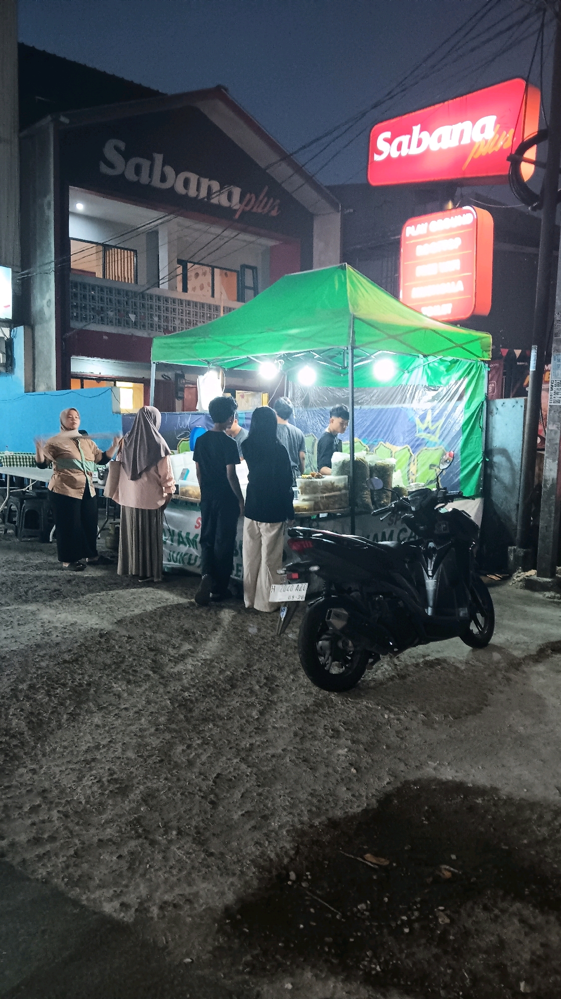 The image captures a street scene at night in South Tangerang, Indonesia, likely near a business named "Sabana Plus," as indicated by signage on a building and a red illuminated sign. In the foreground, a dark-colored motorcycle is parked on a wet, textured pavement. Behind the motorcycle, a green tent is set up, illuminated by overhead lights, suggesting a food stall or market. Several people are gathered around the tent. Two women, one wearing a light brown outfit and a headscarf, and another in a grey headscarf and a long skirt, appear to be interacting with the stall. Three younger individuals, two males and one female, are also present near the tent, with some appearing to be customers or staff. The environment is dimly lit, with artificial lights from the stall and signs illuminating the scene. The building behind the tent has a distinct architectural style with horizontal louvers on the upper floor. The overall atmosphere is that of a bustling night market or street food vendor operation.