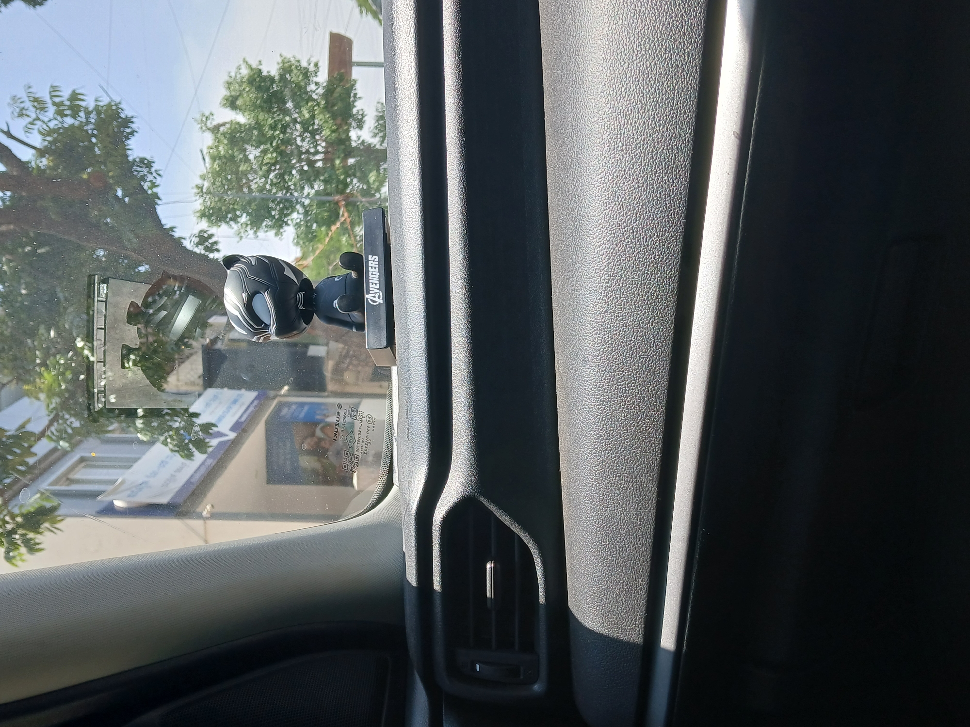 The image depicts the interior of a car, looking out the front window. A Black Panther bobblehead is mounted on the dashboard, with the word "Avengers" printed on its stand. The view through the windshield shows trees and a building with a sign. Based on the QR code visible on the glass it is confirmed to be a SUZUKI Maruti car and the image was taken inside of the car on a sunny day. The building visible through the windshield is a showroom on Bannerghatta Main Rd, Bengaluru, Karnataka, India.