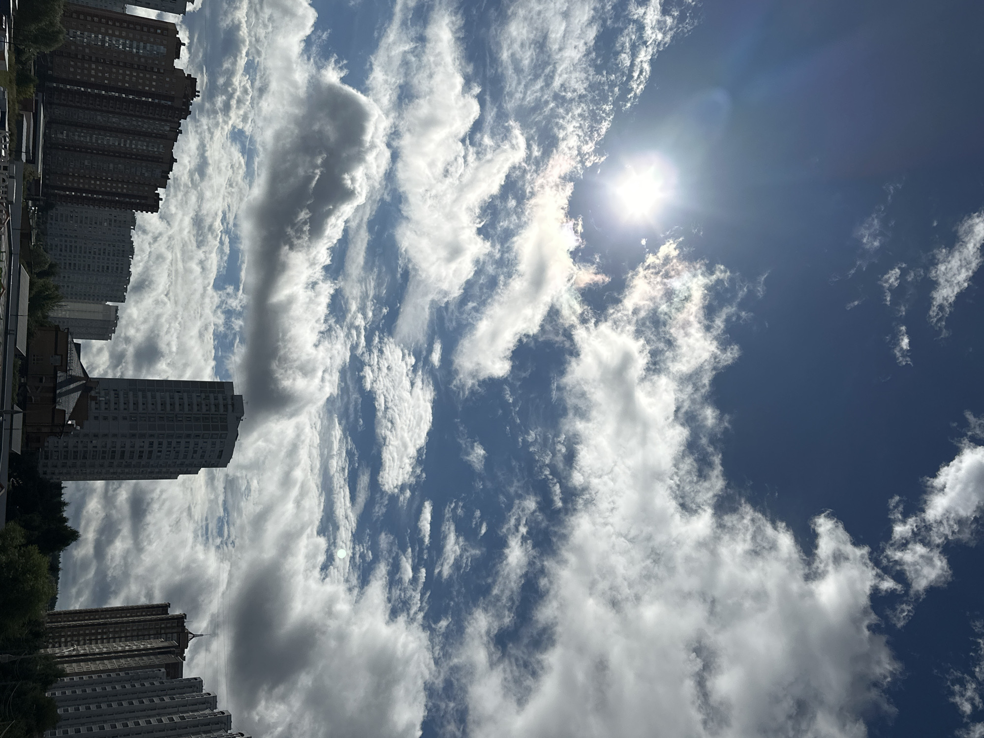 The image shows a view of the city of Podolsk, Russia, on a partly cloudy day. The sun is visible in the upper left corner of the sky, peeking through the clouds, creating a bright glare. The sky is a deep blue, filled with a mixture of white and grey clouds of varying sizes and densities. In the foreground, several tall residential buildings are visible. The building on the far left is a reddish-brown high-rise. Next to it are other multi-story buildings. Towards the center of the image, a taller, lighter-colored building with many windows stands out. To the right, more tall buildings, some appearing to be of modern construction with many windows, are visible. The lower part of the image shows some trees and what appear to be the tops of smaller structures or roofs. The overall scene depicts an urban landscape with a dynamic sky.