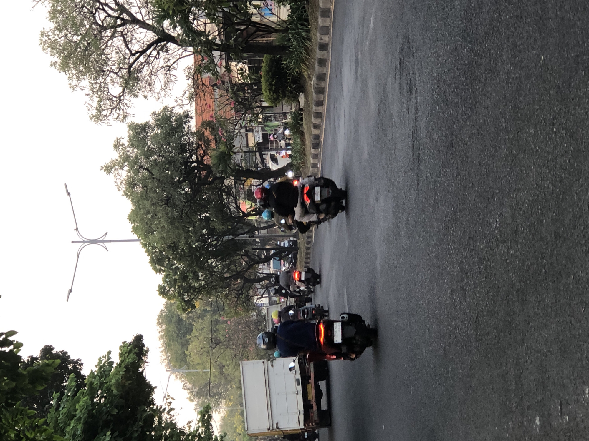 The image depicts a street scene in Sumurame, Indonesia, likely during the late afternoon or early evening given the lighting. The foreground is dominated by an asphalt road with some scuff marks and debris. Several motorcycles are visible, with riders wearing helmets and various casual clothing.
In the left foreground, a dark-colored scooter with a rider in a dark blue shirt and a grey helmet is moving away from the camera. To the right of this scooter, another scooter is visible from the rear, with a rider wearing a black jacket and a red helmet, also moving away. Further back, on the right side of the road, a line of vehicles, primarily motorcycles, are moving in the same direction.
On the left side of the road, a white truck with a large cargo box is parked or moving slowly. Along the sides of the road, there are numerous trees with dense foliage, some of which overhang the street. In the background, buildings with red tiled roofs and some signage are visible, partially obscured by trees. Streetlights are also present, with one prominent pole in the upper center of the image. The overall impression is a typical urban street with moderate traffic.