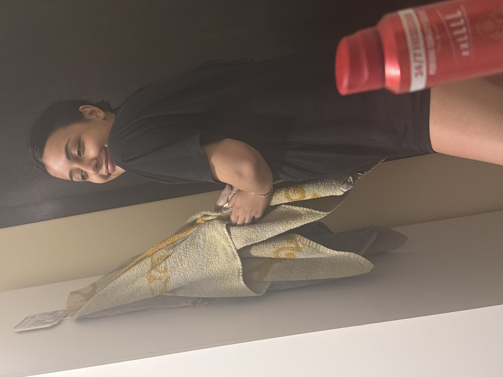 The image, originally rotated 90 degrees clockwise, depicts a young woman smiling at the camera in an indoor setting. She is positioned on the left side of the frame, wearing a black t-shirt and dark shorts. Her expression is cheerful and friendly.
Her hands are engaged with a light-colored, possibly beige or yellow, patterned towel resting on a white shelf or surface. The towel has a recurring dark pattern. Partially visible beneath the towel, to its left, is a small, flat white object.
In the upper right portion of the image, a red spray bottle or can is visible, appearing to be held or placed nearby. Text on the bottle includes "24/7 FRESH" and "111", and part of a brand name that appears to be "AXE".
The background behind the woman is dark, possibly a wall or dark furniture. Below the towel and shelf, there is a prominent white surface, and above the shelf, a section of light beige wall with a dark horizontal line, possibly a trim or another shelf edge. There are no specific visual cues within the image to indicate the provided location context of Shumen, Bulgaria.