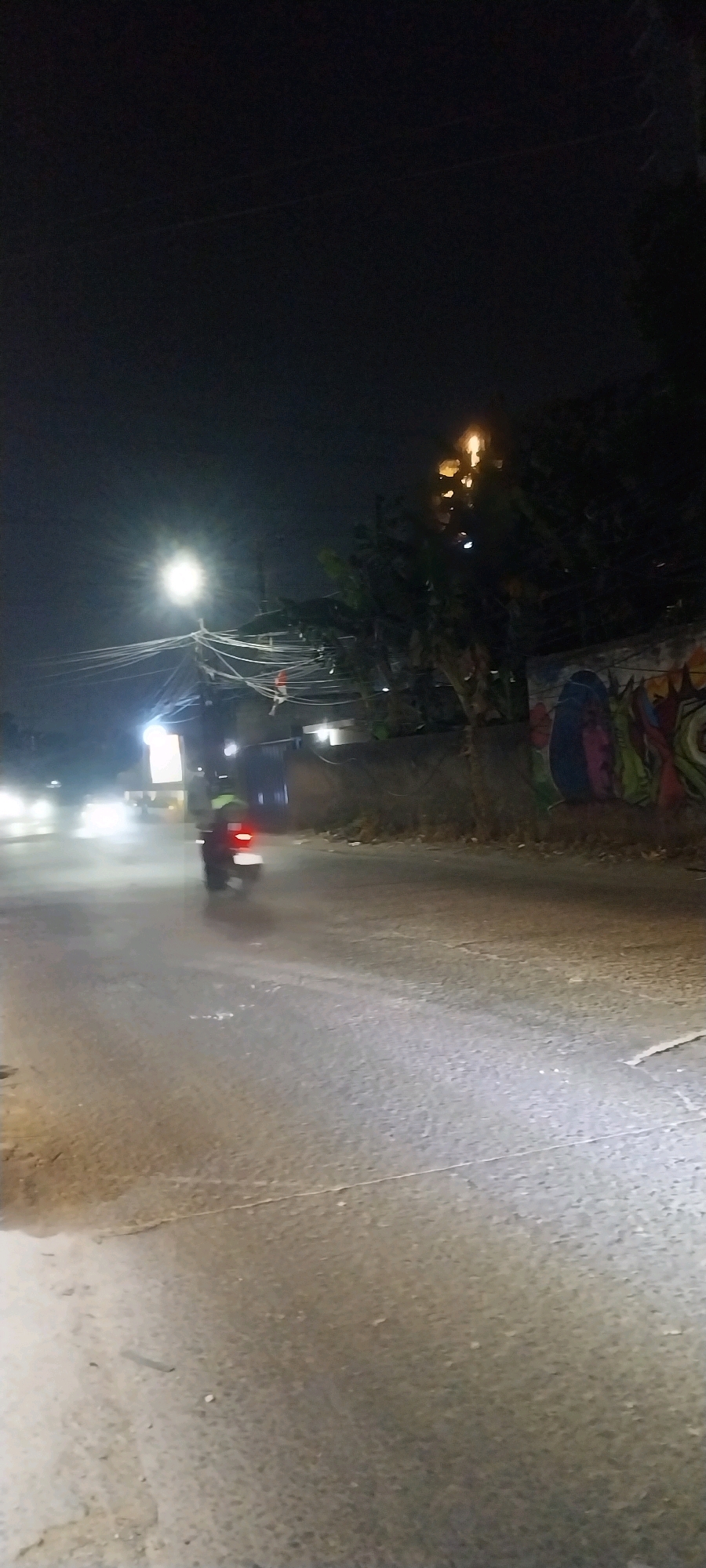 The image depicts a nighttime street scene in Bojong Nangka, Indonesia. A motorcycle with its brake light illuminated is moving away from the camera on a wet, asphalt road. The road surface appears worn and shows cracks. On the right side of the road, a graffiti-covered wall is visible, with trees and vegetation behind it. Overhead, a dense network of electrical wires crisscrosses the sky, illuminated by a bright street light. In the distance, blurred headlights of other vehicles can be seen, suggesting ongoing traffic. The overall atmosphere is dark, with the artificial lighting creating highlights and shadows on the street.