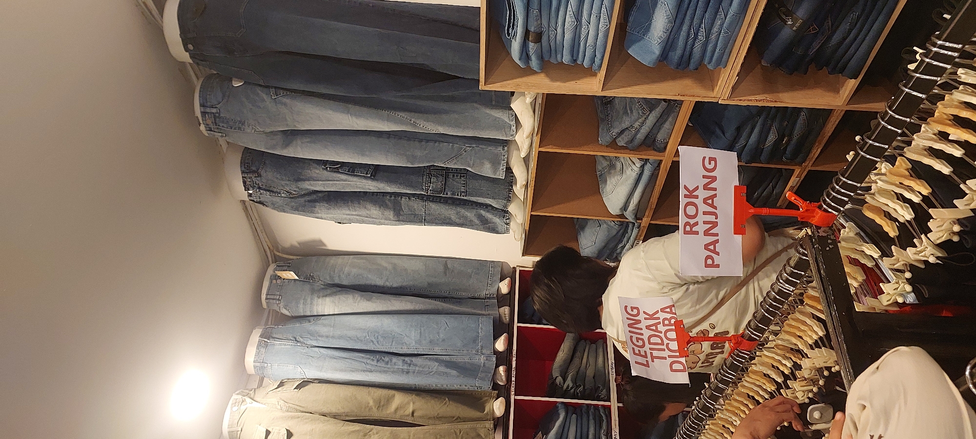 The image depicts the interior of a clothing store. In the foreground, several racks of clothing are visible, with a person in a light-colored t-shirt standing in front of a shelf filled with folded jeans. Two signs are prominently displayed: one reads "LEGING TIDAK DICOBA" (Leggings not to be tried on), and the other reads "ROK PANJANG" (Long skirt). In the background, mannequins dressed in various styles of jeans are displayed on a raised platform. The store appears to be well-lit. This scene is likely located in Jayapura, Indonesia, given the Indonesian language on the signs.