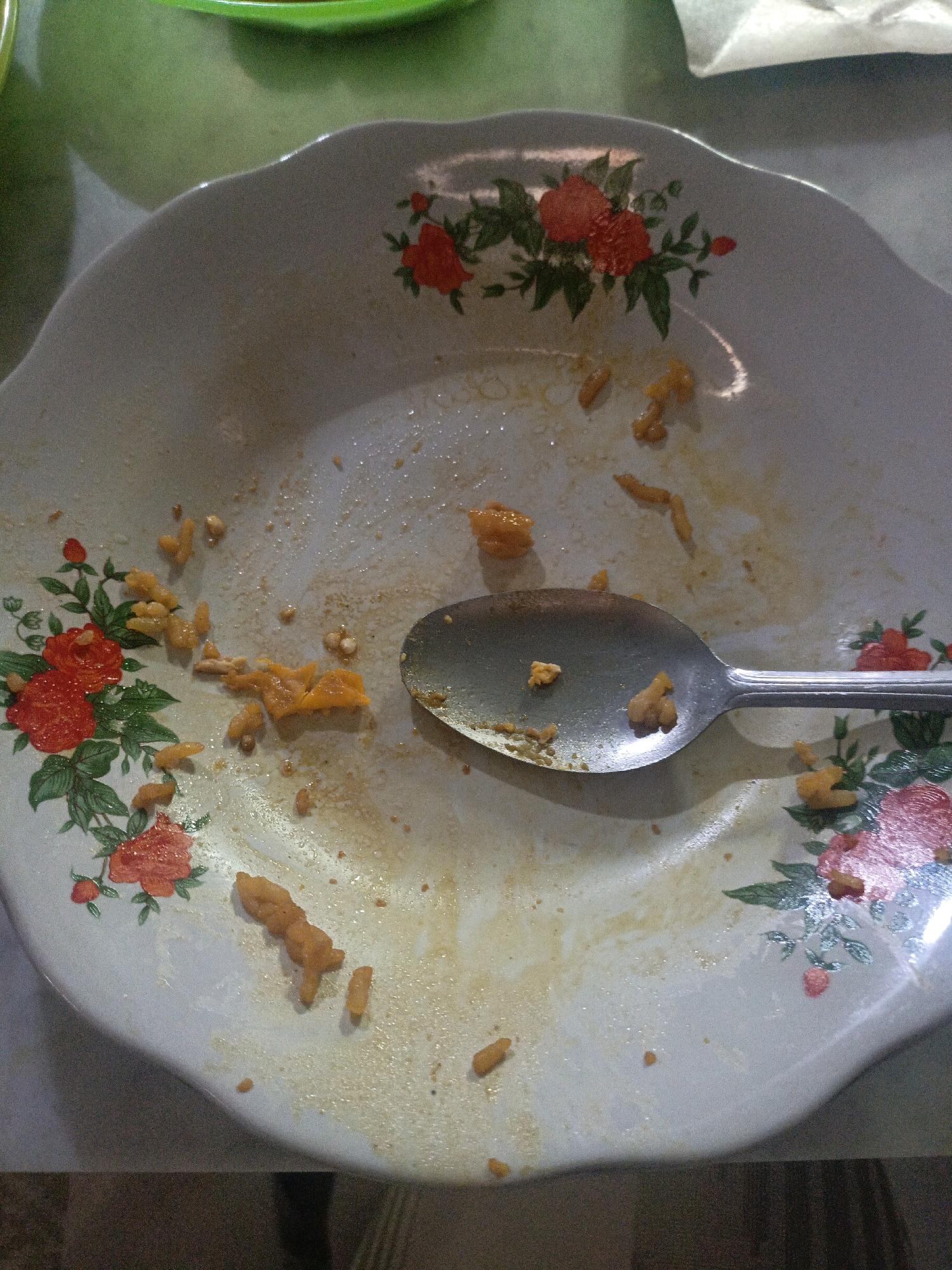 A close-up, high-angle shot shows the remnants of a meal on a floral-patterned plate. The plate, white with red and pink flowers and green leaves, is mostly empty, with scattered yellow pieces of food and sauce residue. A metal spoon rests in the lower right quadrant of the plate, also with some food particles on it. The background is blurred, with hints of green and white suggesting other objects or surfaces. The image appears to be taken indoors, likely in a domestic setting in Undaan, Indonesia, with the lighting suggesting an indoor environment. There are no people visible in the frame.