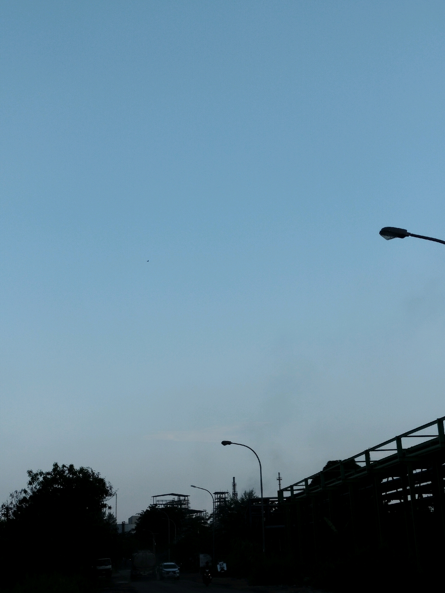 The image shows a silhouetted industrial landscape against a pale blue sky. In the foreground, a dark green elevated structure with a railing dominates the right side of the frame. A lamppost is visible on the left side of this structure, and another lamppost is positioned further back on the left side of the image. To the left, the silhouettes of trees and a few vehicles, including a truck and a car, are present. In the background, industrial buildings with towers and piping are visible. A small dark object, possibly a bird or an insect, is discernible in the upper left quadrant of the sky. The scene appears to be captured during dusk or dawn, given the lighting conditions. The location is identified as a road near an industrial area in Batam, Indonesia.