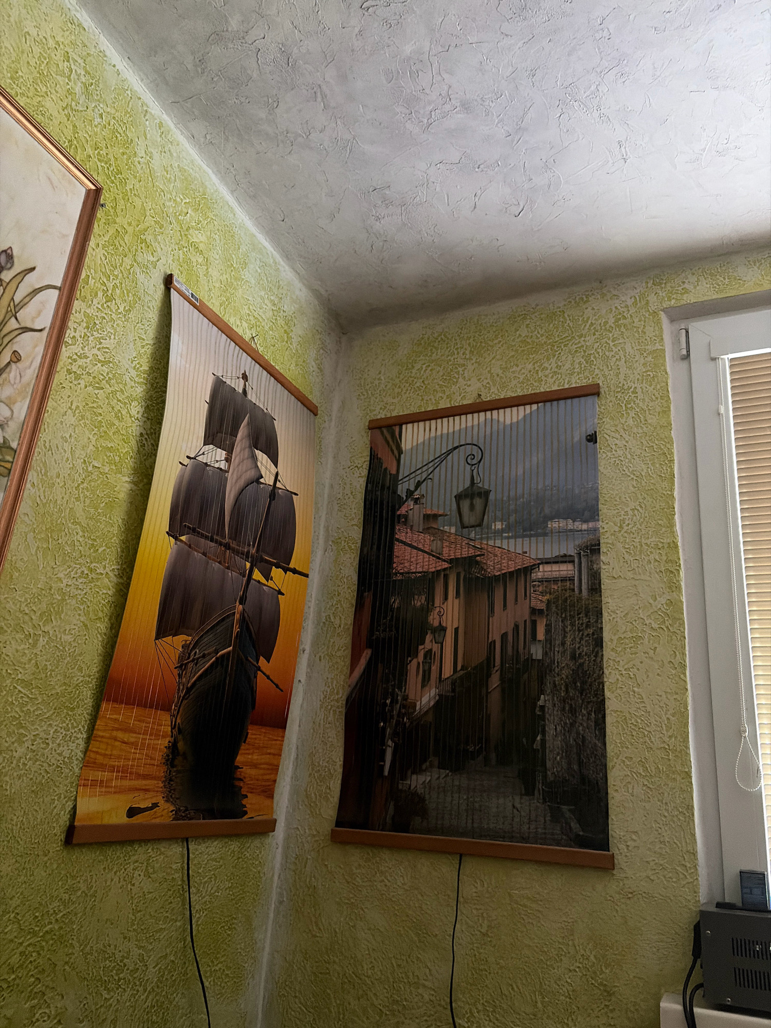 The image shows an interior corner of a room with textured light green walls and a textured white ceiling. Two framed artworks are hung on the wall. The artwork on the left is a depiction of a tall sailing ship at sunset, with its sails full and reflected in the water. The artwork on the right depicts a European street scene with old buildings and a lamppost, leading towards a body of water with buildings in the distance. A window with blinds is visible on the right side of the image, and some electronic equipment is placed on a surface below the window. The artworks appear to be printed on a material that is stretched over wooden frames. There are no people visible in the image. The overall impression is of a residential interior. The metadata suggests the location is Ливадийский поселковый совет, Ukraine.