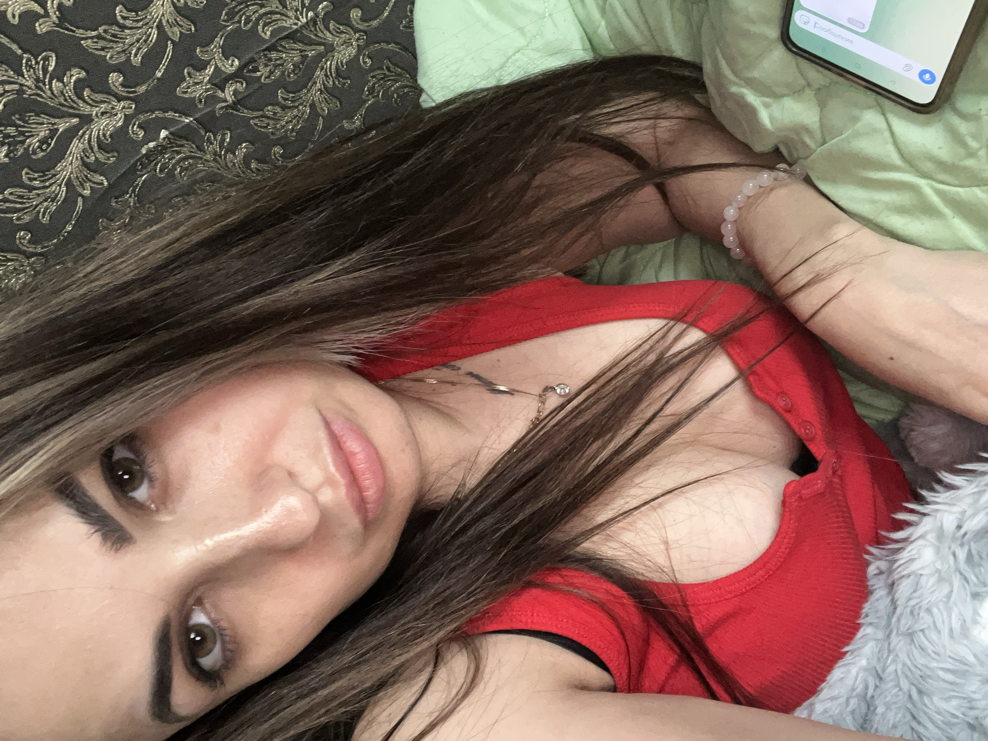 A woman with long, dark brown hair lies comfortably, captured in a slightly elevated, close-up shot that accentuates her facial features and upper body. She is wearing a red top with visible buttons down the front. A delicate gold chain with a small pendant adorns her neck. On her right wrist, she wears a bracelet made of light-colored, possibly rose quartz, beads.
Her gaze is directed towards the camera, with her brown eyes appearing to make direct contact. Her lips are slightly parted, revealing a natural, perhaps neutral, expression. Her eyebrows are dark and well-defined, and she has visible eyeliner on her eyelids.
The setting suggests an indoor environment, likely a bedroom or living space, given the soft textures and patterned fabric seen in the background. To the upper left, a dark fabric with a gold, ornate floral or vine pattern is visible, possibly part of a pillow or headboard. On the right side, a light green textured fabric, perhaps a sheet or blanket, is present, with some loose threads. Further to the right and slightly above her head, a smartphone is visible, resting on the green fabric. The phone screen shows what appears to be a messaging application interface, with some faint text in Russian visible: "Сообщение" (Message) and possibly "Голосовое" (Voice). A microphone icon is also discernible on the screen. A fluffy, light grey material is also partially visible on the bottom right, possibly another blanket or cushion.
The lighting appears soft and even, suggesting indoor artificial light or diffuse natural light. The overall mood is relaxed and intimate.