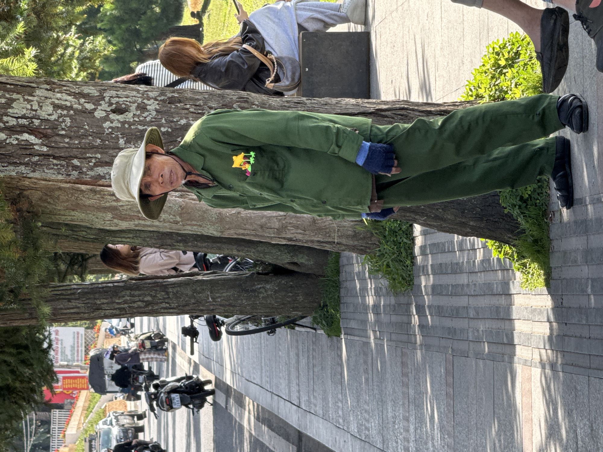 An elderly Vietnamese man wearing a green uniform and a wide-brimmed hat stands on a sidewalk in Da Lat, Vietnam. He has dark hair and a weathered face. His uniform has a colorful patch with yellow and green figures on the left breast pocket. He wears blue fingerless gloves and dark sandals. Behind him, a woman with auburn hair, wearing a black jacket and grey sweatpants, sits on a black bench and uses a mobile phone. The sidewalk is paved with grey and pinkish rectangular tiles. Several trees line the sidewalk, casting shadows on the ground. In the background, a street is visible with motorcycles and cars.