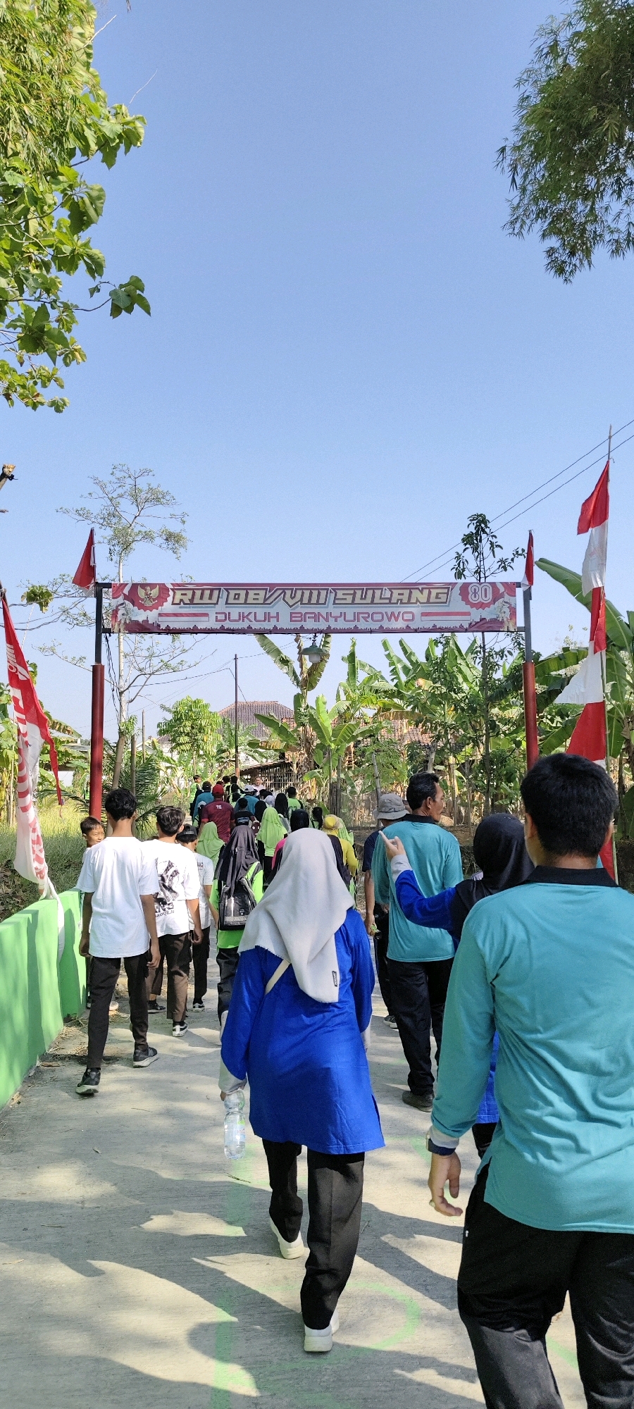 The image depicts a group of people walking down a paved path lined with a green fence on the left and banana trees and other vegetation on the right. They are walking under a banner that reads "RW VIII SULANG DUKUH BANYUROYO 80" and is adorned with Indonesian flags. The sky is clear and blue, suggesting it is daytime. The people appear to be participating in an outdoor activity, possibly a procession or a walk for a local event. The overall atmosphere is communal and festive, with the prominent display of national symbols indicating a civic celebration. The location is identified as RW VIII Sulang, Dukuh Banyuroyo, which is in the Sulang sub-district, Rembang Regency, Central Java, Indonesia.