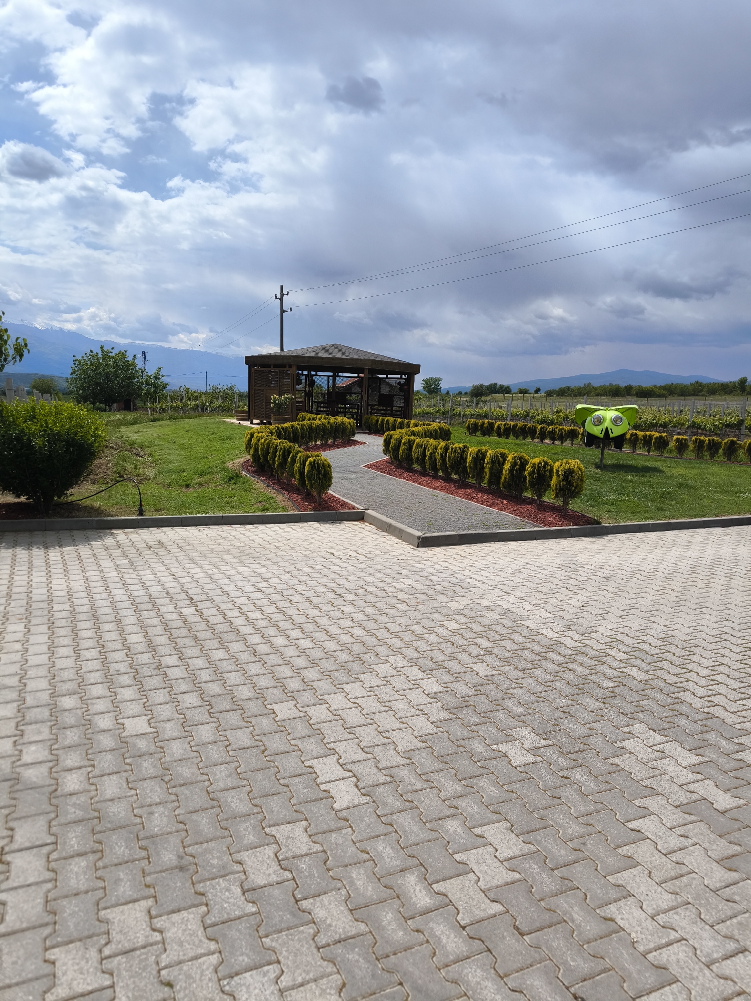 The image shows a gazebo in Kapatovo, Bulgaria. The gazebo is constructed from dark wood and is located on a green lawn. In front of the gazebo, there is a gravel path bordered by small, neatly trimmed bushes and red mulch. A paved area composed of gray interlocking blocks is in the foreground. Further behind the gazebo and bushes, there are rows of vineyards. In the distance, there are mountains under a sky with partly cloudy conditions. A green decorative structure is also present near the gazebo. Overhead power lines are visible in the sky.